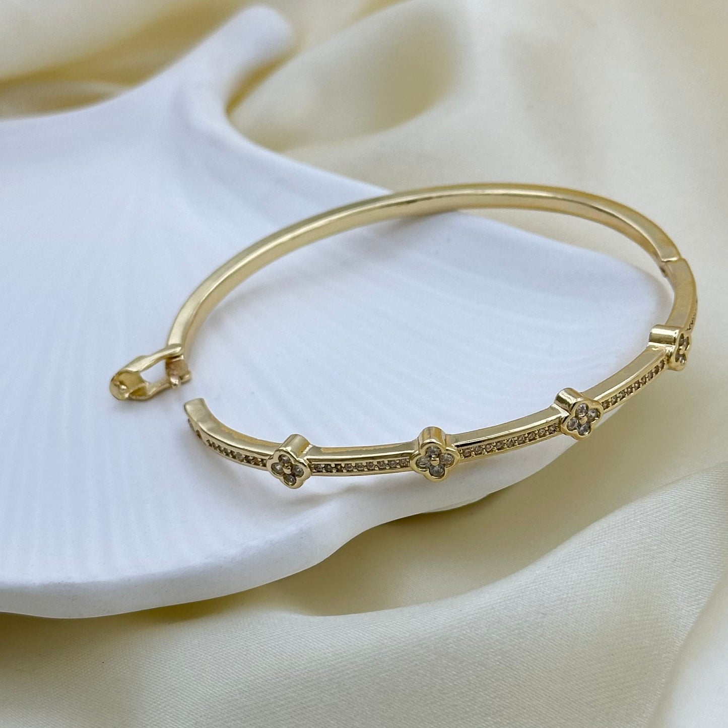 A gold-plated bangle bracelet with small clear crystal accents, displayed on a soft white textured surface with draped cream fabric in the background.