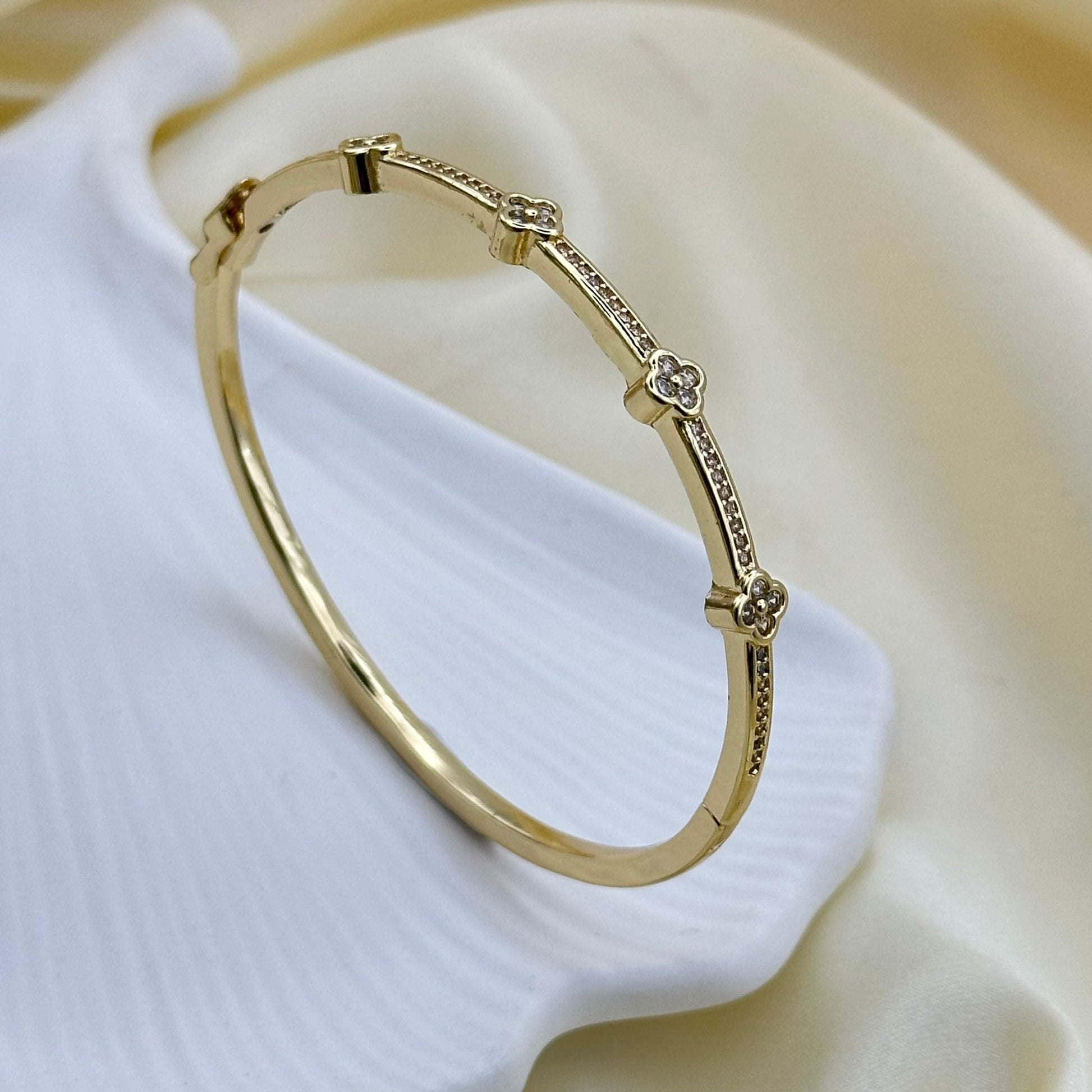 A gold bangle bracelet with small floral motifs and textured details, displayed on a soft white fabric background.