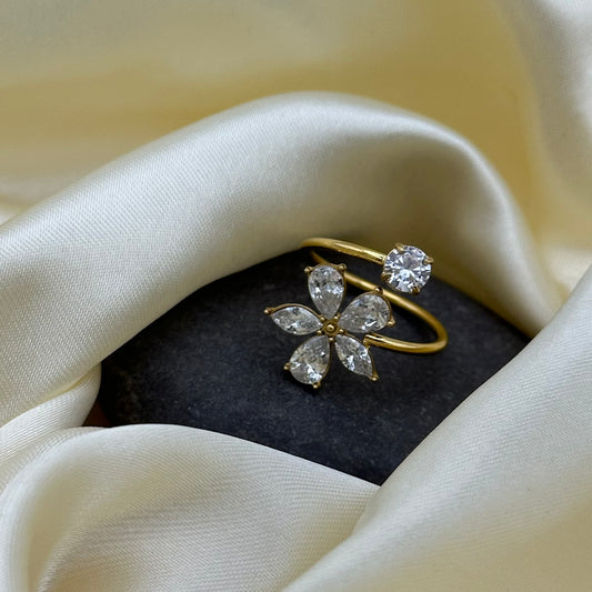 A gold flower-shaped ring with clear gemstones sits on a dark stone surrounded by draped cream satin fabric.