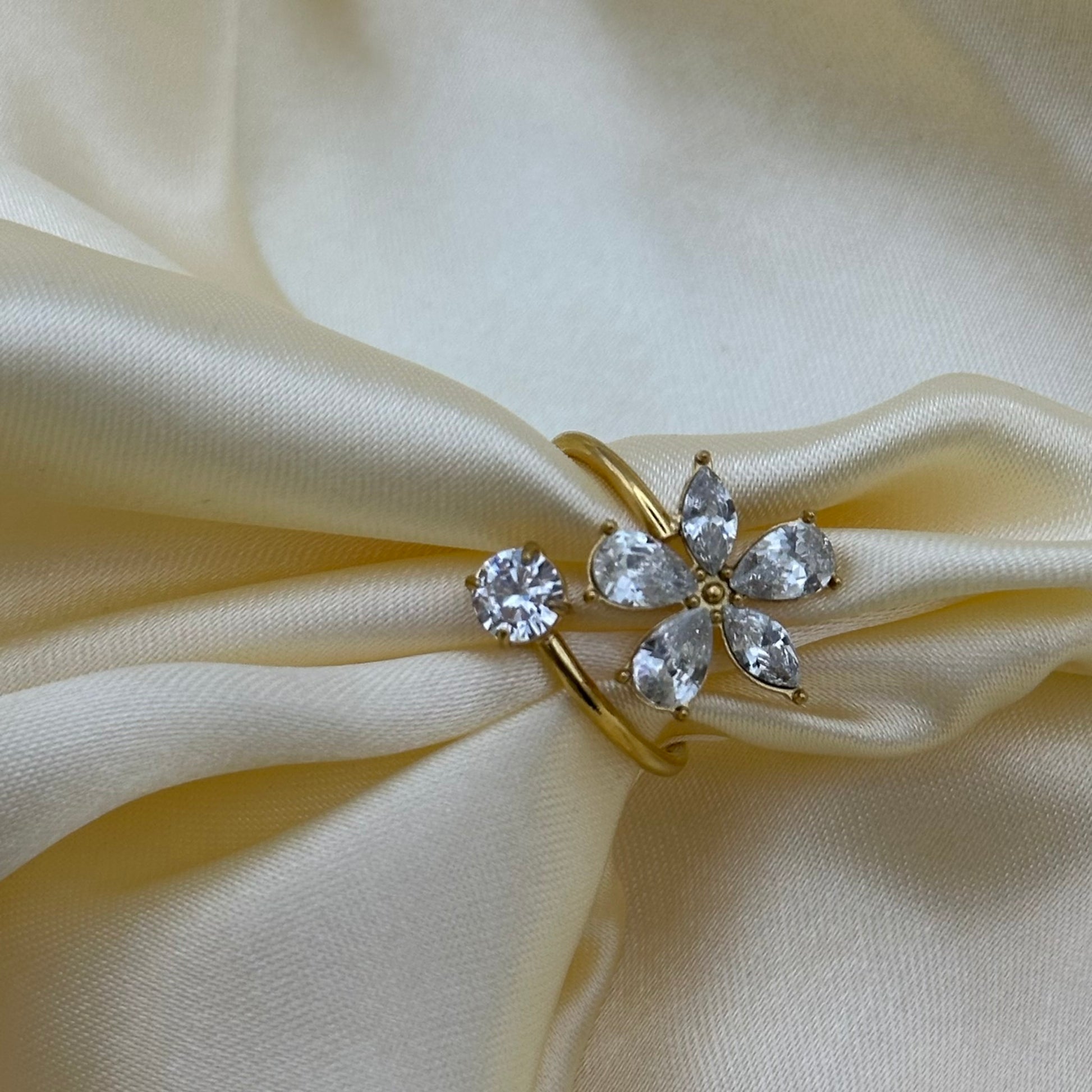 A gold ring featuring a flower-shaped cluster of clear teardrop and round gemstones, displayed on draped cream satin fabric.