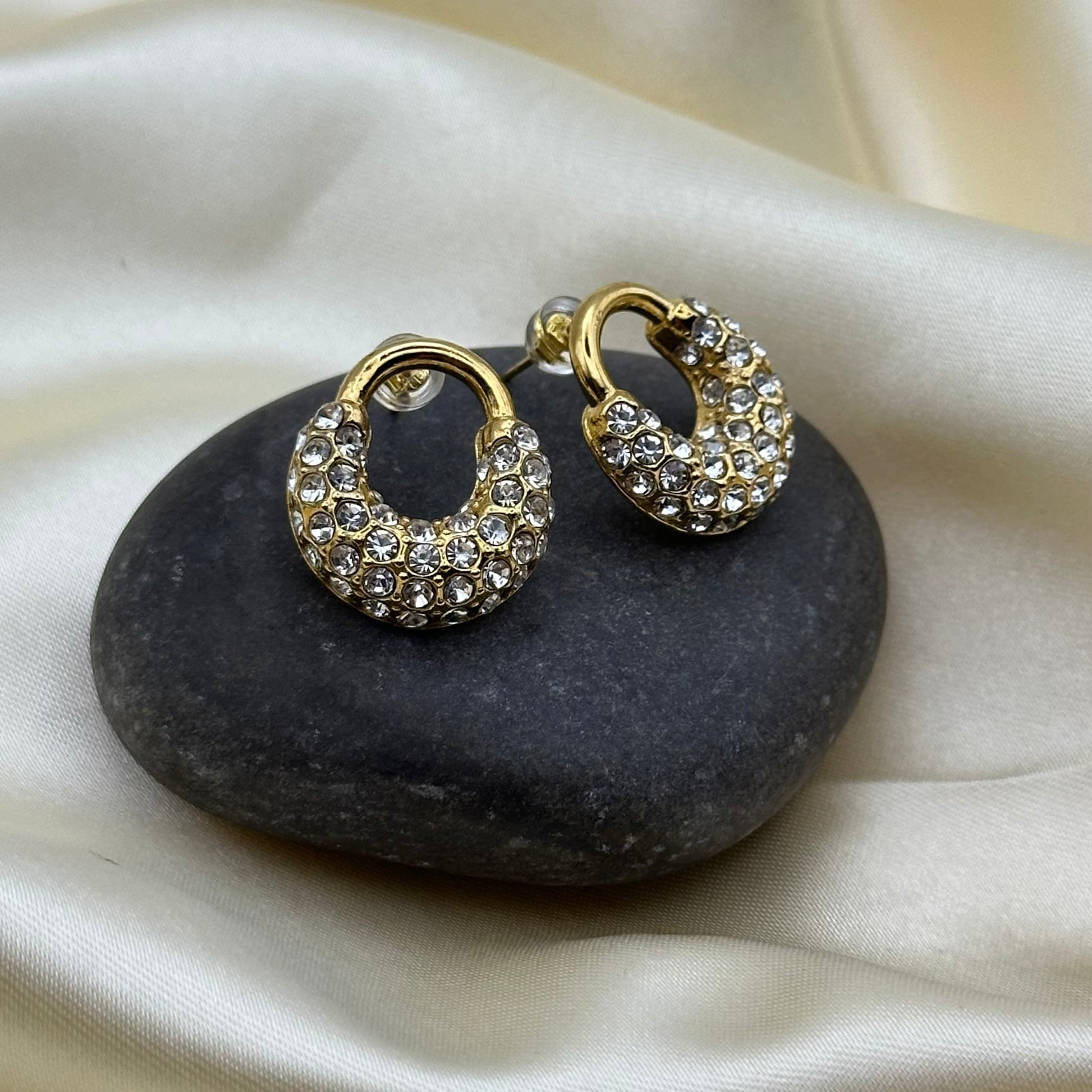 A pair of gold crescent-shaped hoop earrings covered in clear rhinestones, displayed on a dark stone over a cream satin fabric background.