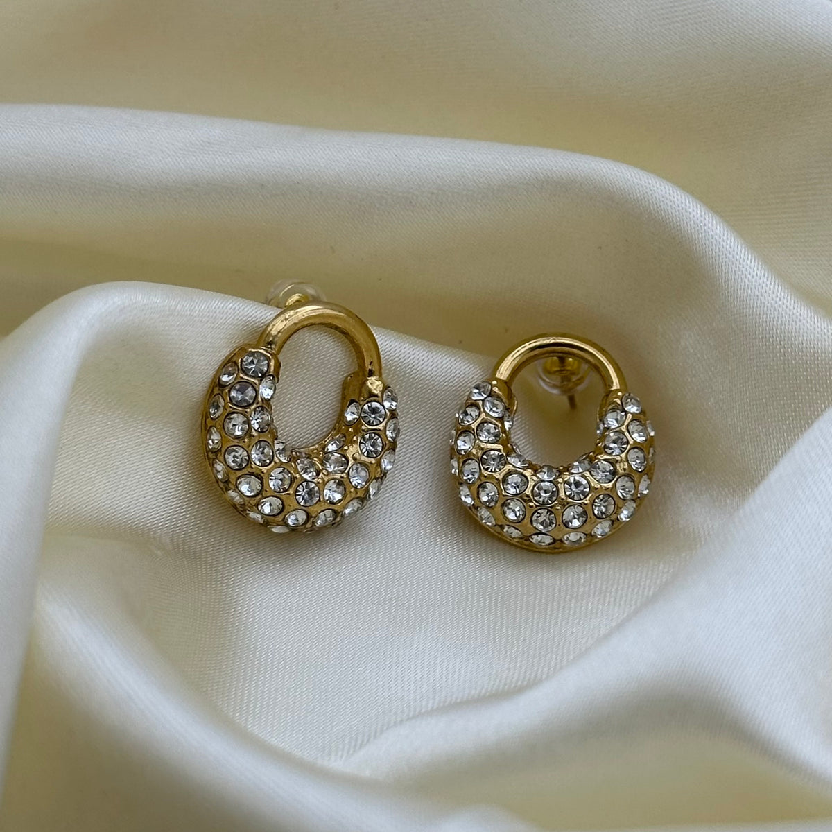 A pair of gold crescent-shaped hoop earrings covered in clear rhinestones, displayed on draped cream-colored satin fabric.