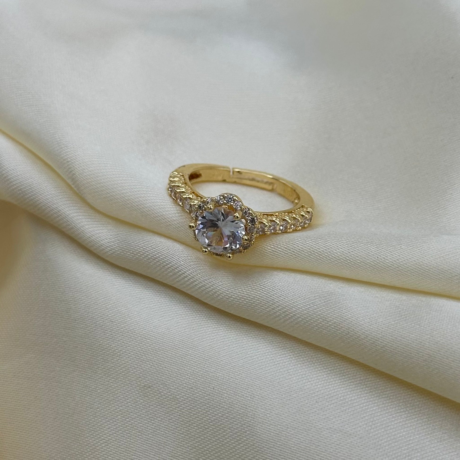 A gold ring with a heart-shaped clear gemstone and small accent stones, displayed on draped cream-colored satin fabric.