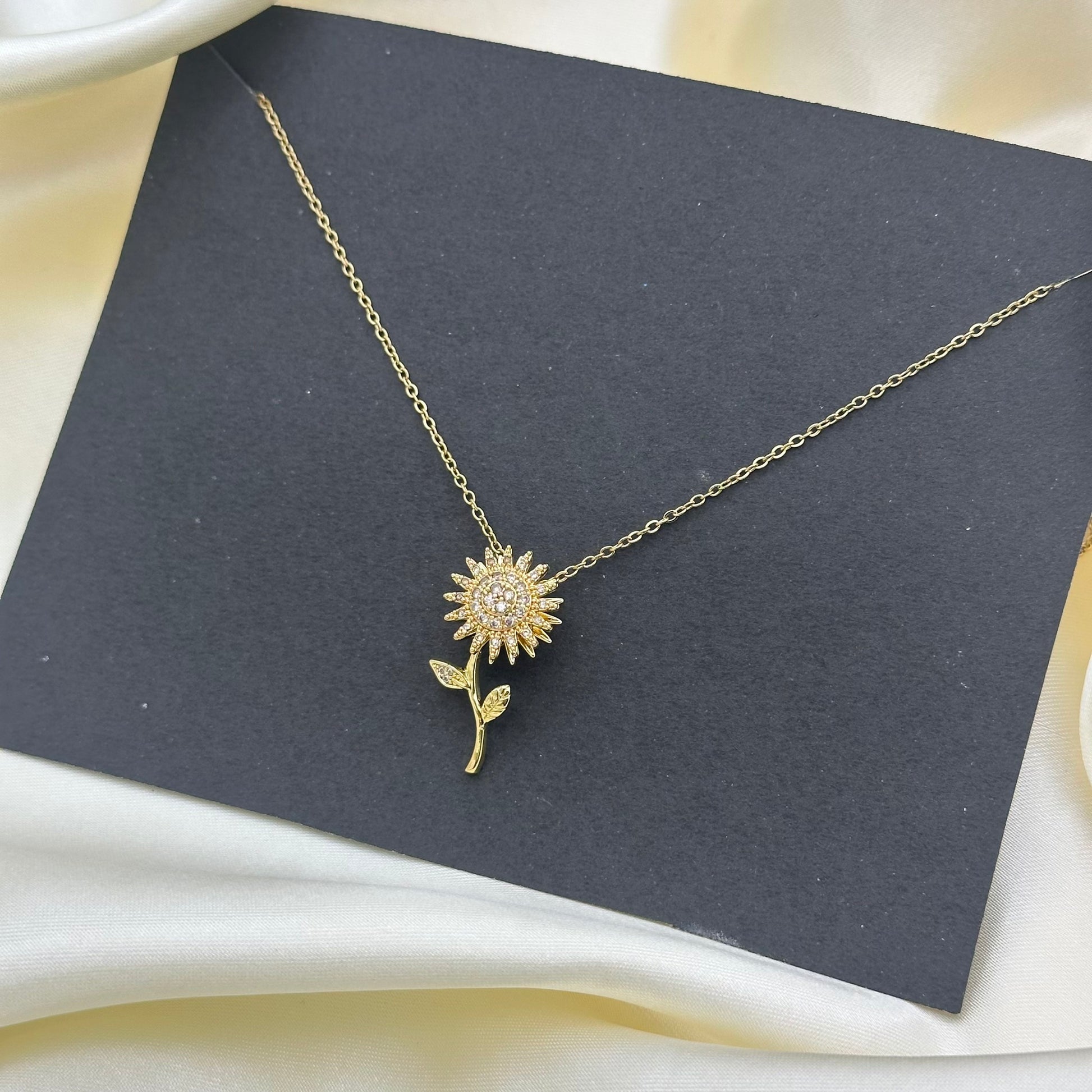 A gold necklace with a flower pendant, featuring clear rhinestones in the center, displayed on a dark gray card over a cream satin fabric background.