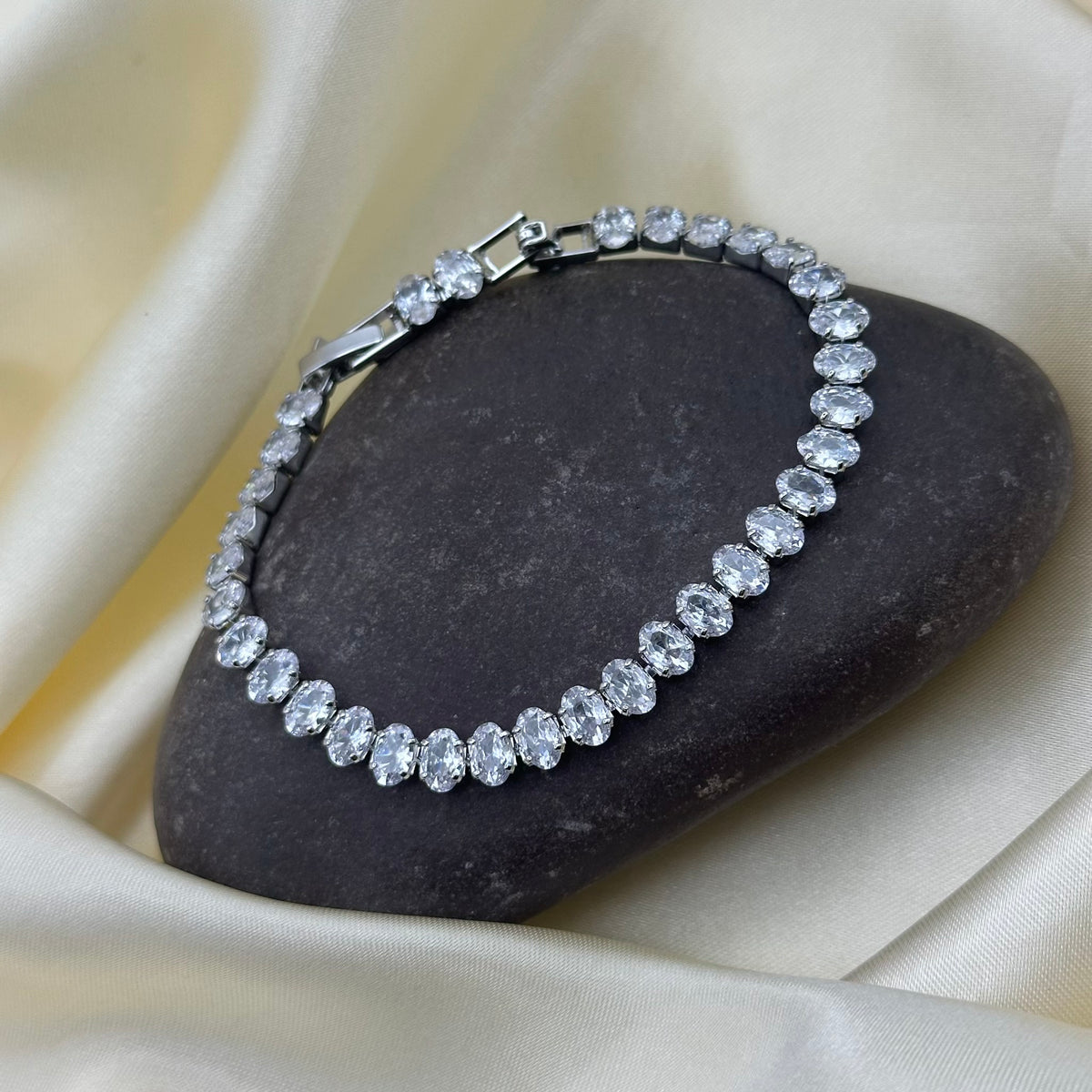 A silver tennis bracelet with oval clear stones, displayed on a dark stone over draped cream satin fabric.