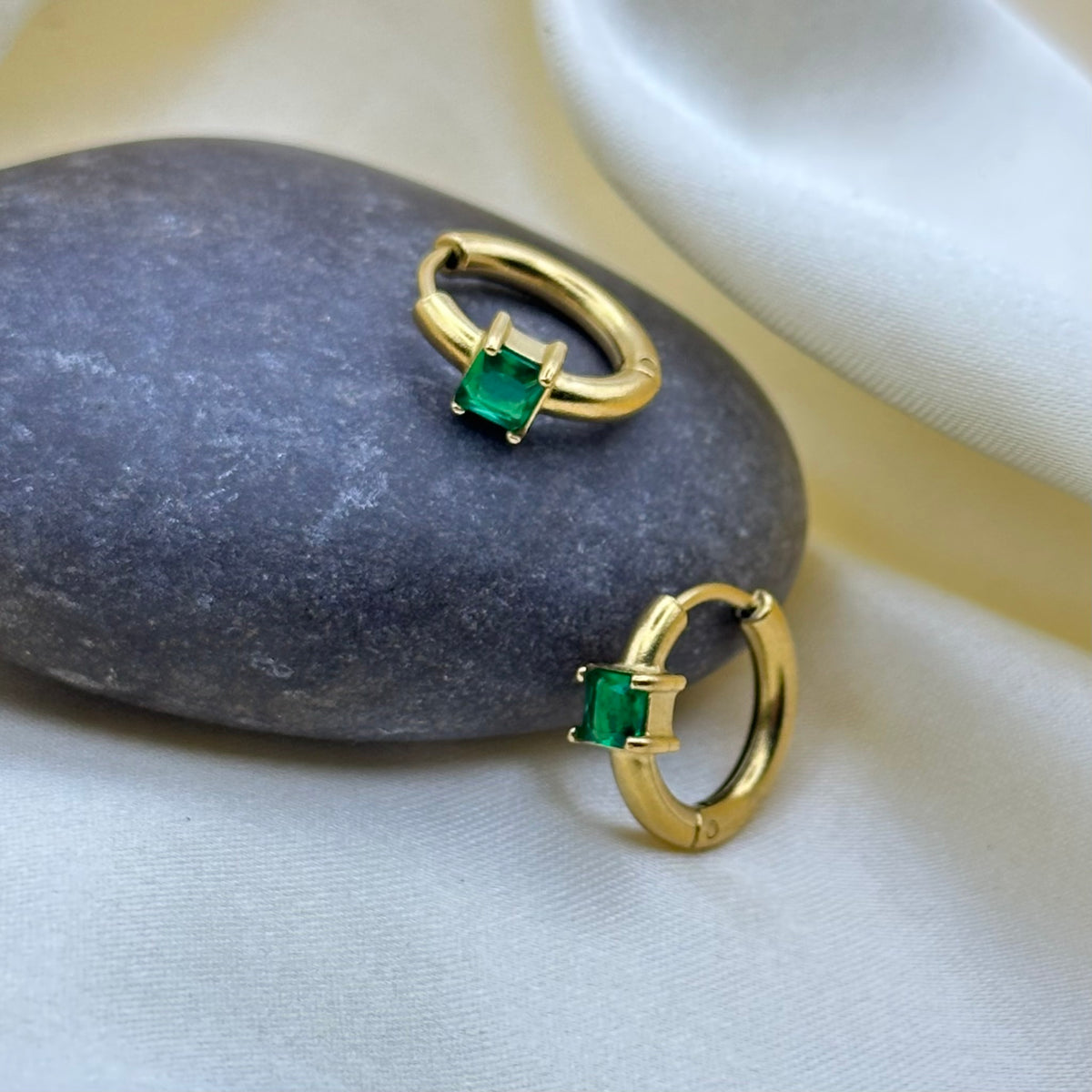 A pair of gold huggie hoop earrings with square-cut green emerald gemstones, displayed on a dark gray stone and white fabric.