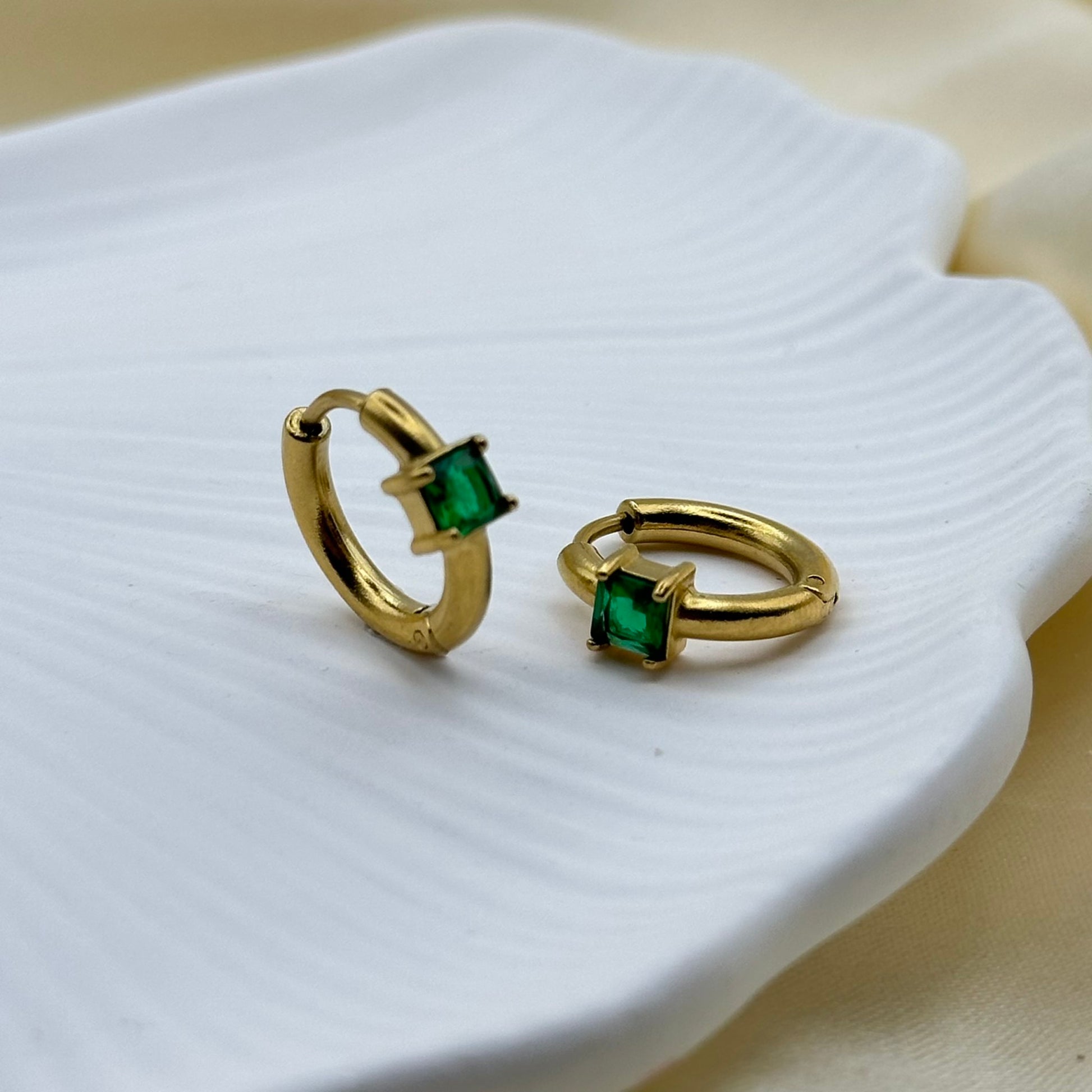 A pair of gold huggie hoop earrings with square-cut green gemstones, displayed on a white ceramic dish over a soft beige fabric background.