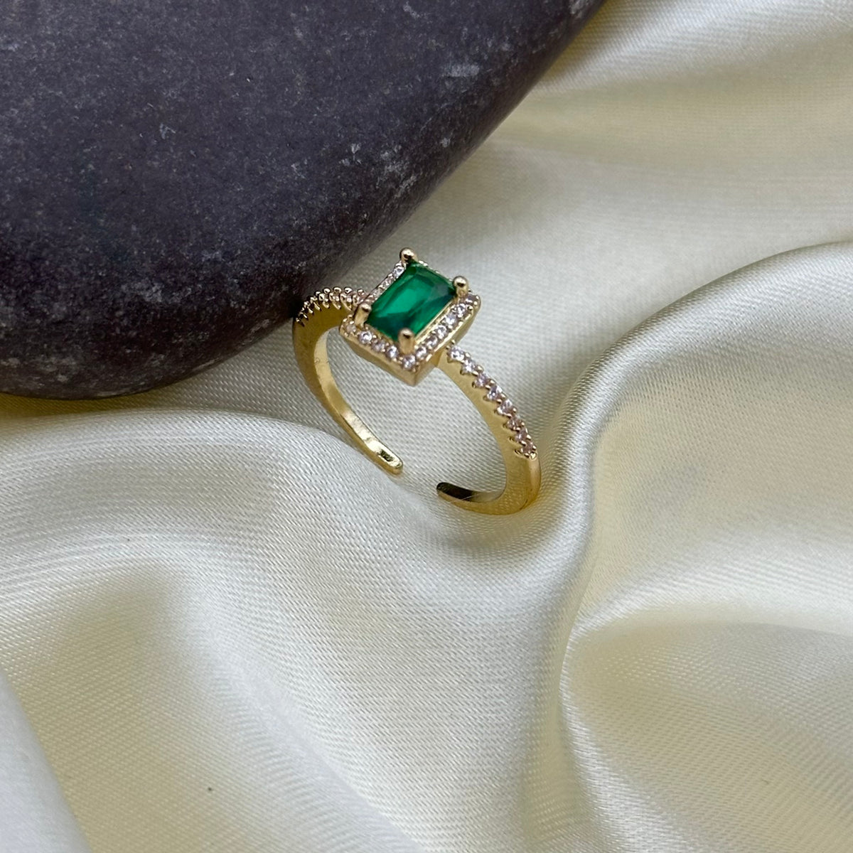 A gold-plated open ring with a rectangular green emerald-cut gemstone and small clear accent stones, displayed on draped cream satin fabric next to a dark stone.