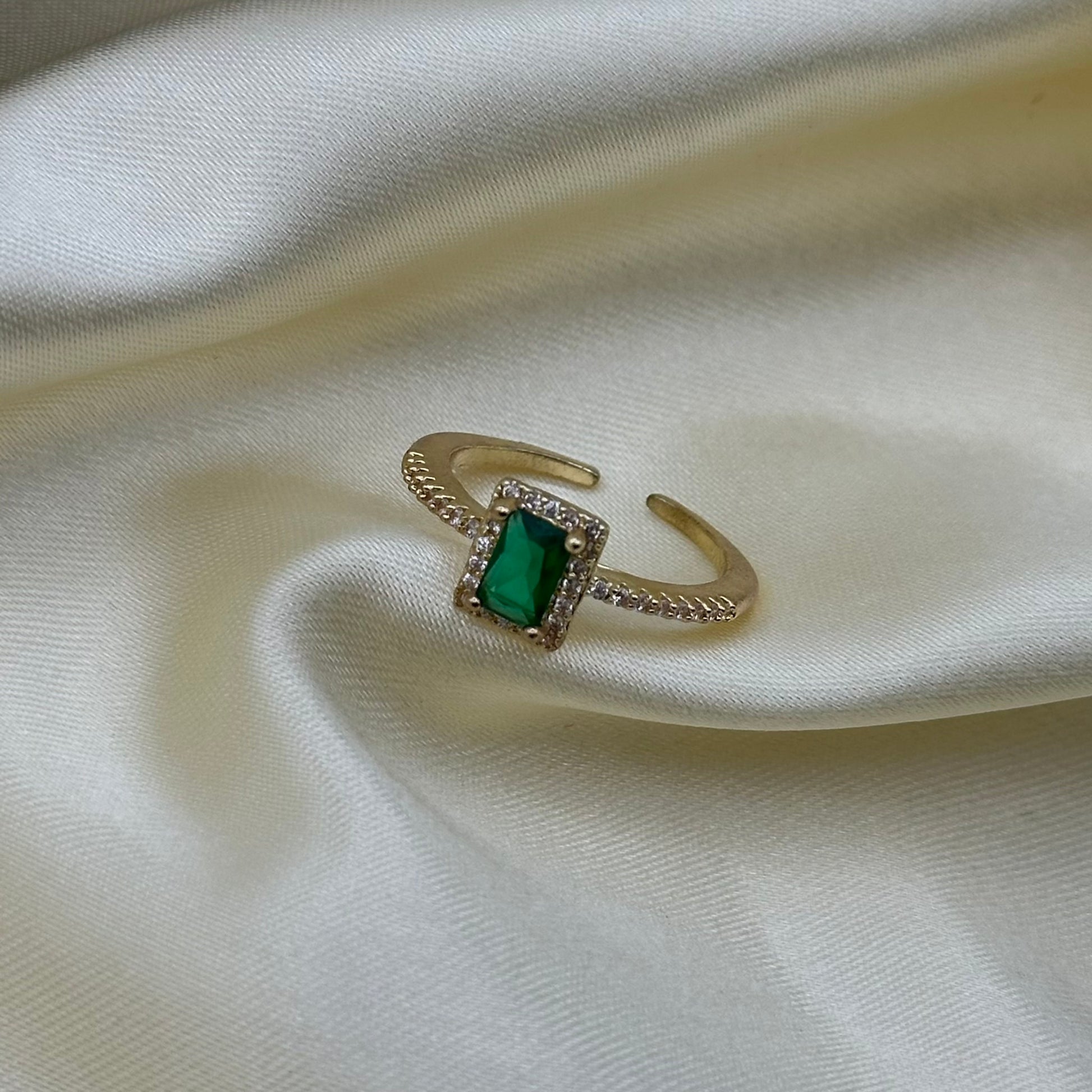 A gold-toned adjustable ring featuring a rectangular green gemstone surrounded by small clear stones, displayed on draped cream satin fabric.