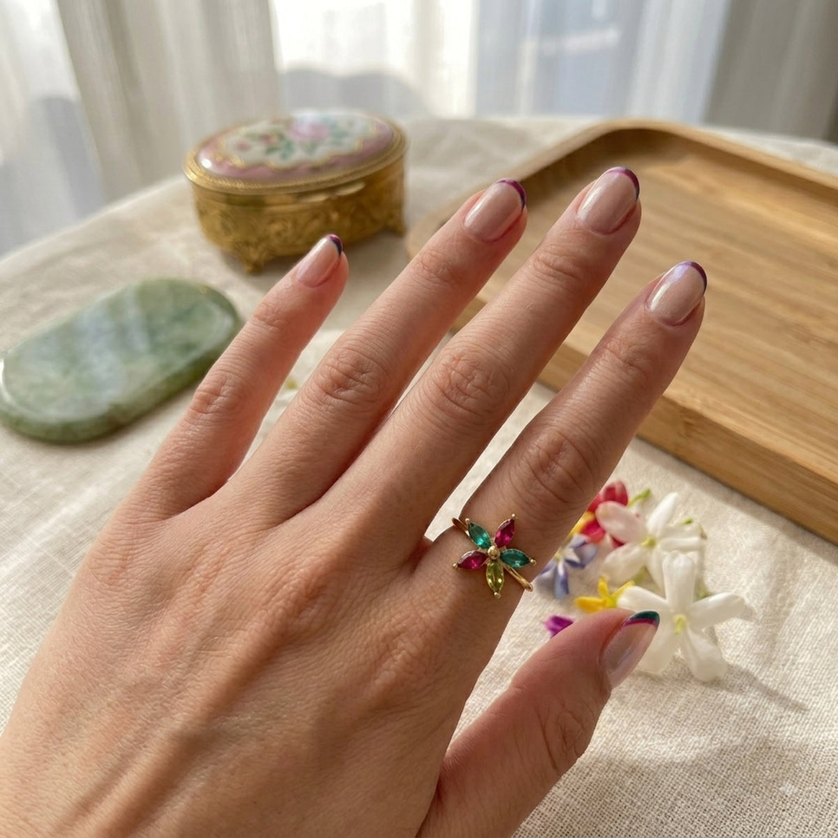 A hand wearing a gold ring with a multicoloured crystal flower, photographed beside jasmine flowers and a vintage trinket box.