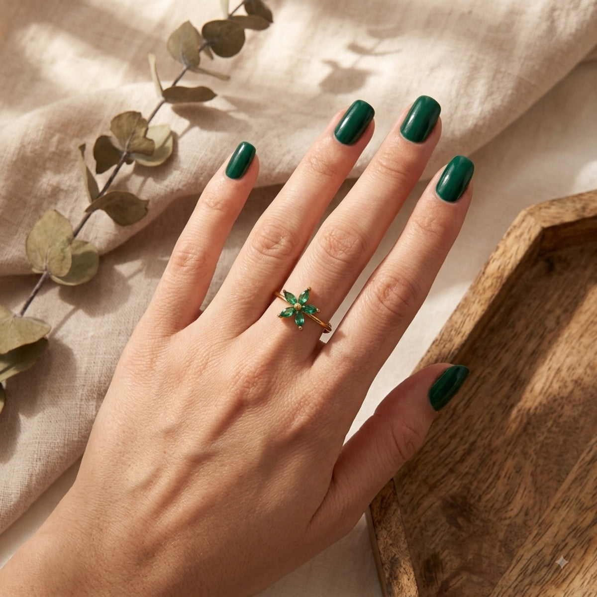 A hand with dark green nails wearing a gold ring with a green crystal flower, styled with dried eucalyptus and a wooden tray.