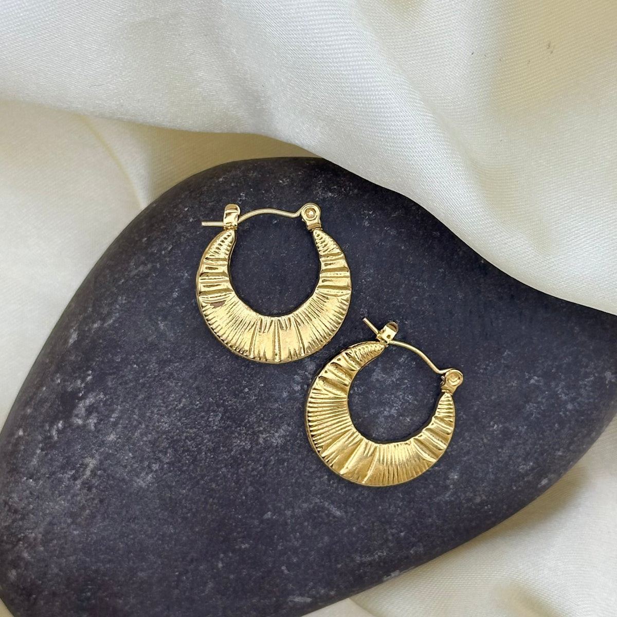 A pair of ribbed gold crescent hoop earrings resting on a dark stone over cream satin fabric.