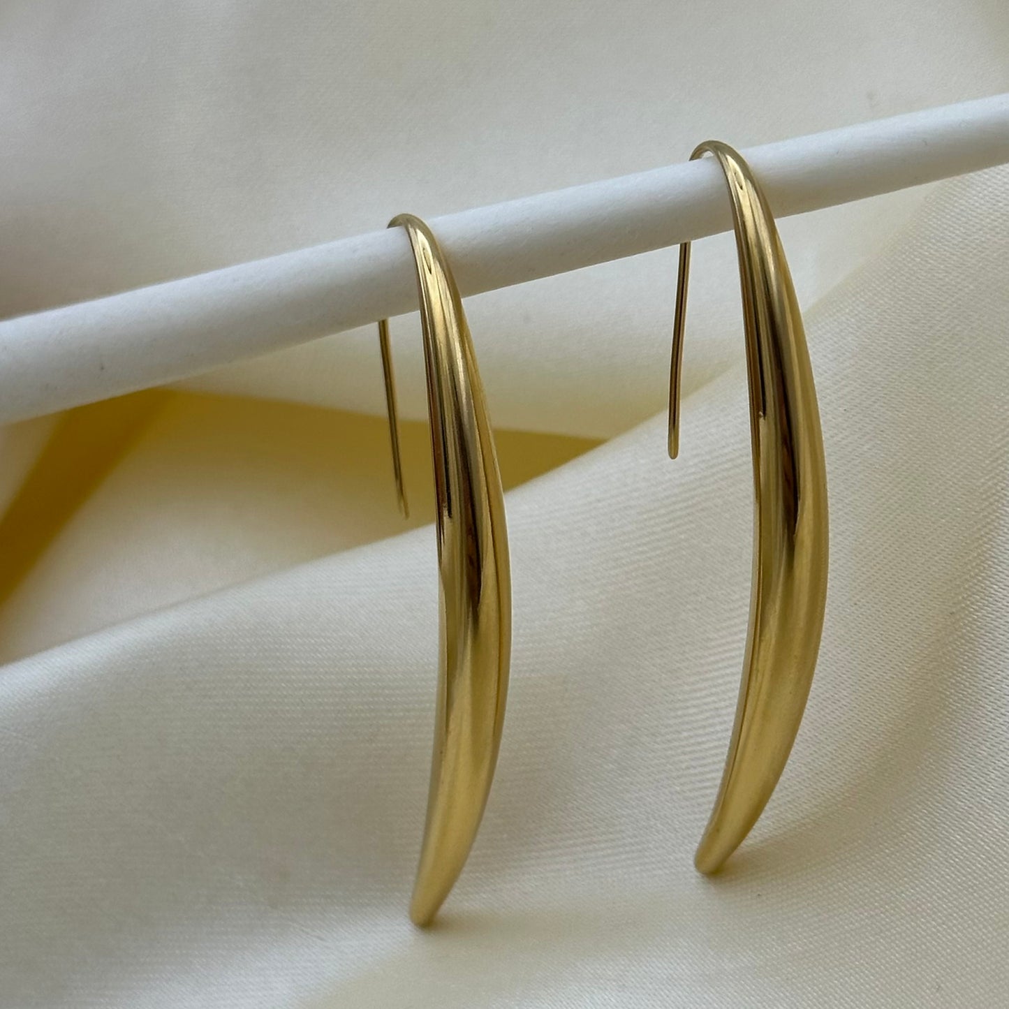 A pair of polished gold crescent-shaped hoop earrings hanging from a white display stand against a soft cream fabric background.