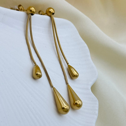A pair of gold dangle earrings with teardrop-shaped pendants, displayed on a white textured surface with a soft beige fabric background.