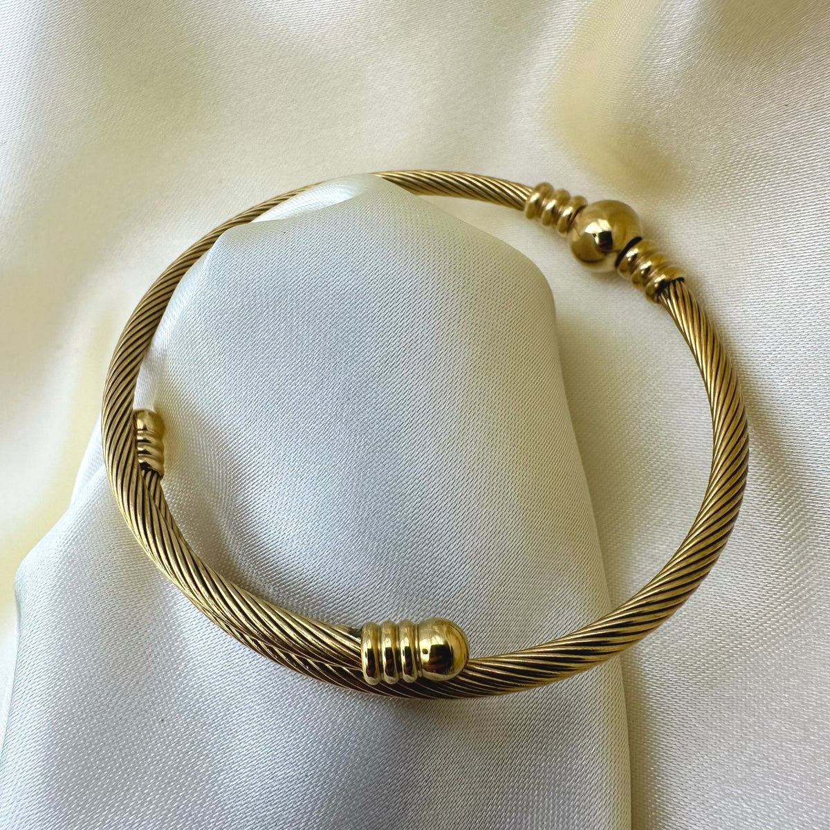 A gold-toned twisted cable bangle bracelet with small bead accents, displayed on a soft white fabric surface.