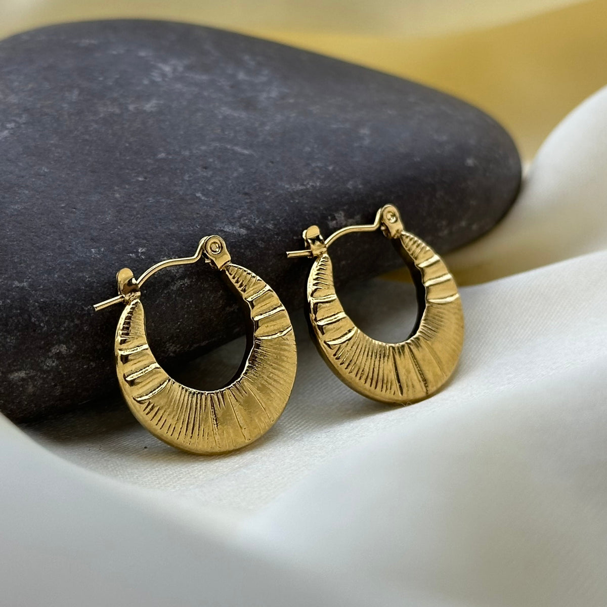 A pair of textured gold hoop earrings resting on a dark stone and white fabric.