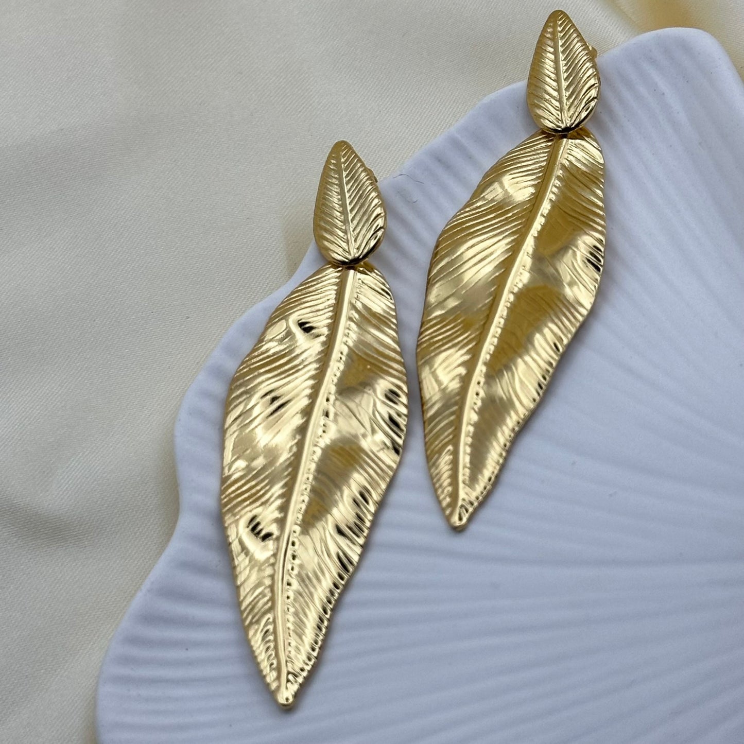 A pair of large gold-toned feather drop earrings with a textured, ribbed surface, displayed on a white ribbed dish over a light beige fabric background.