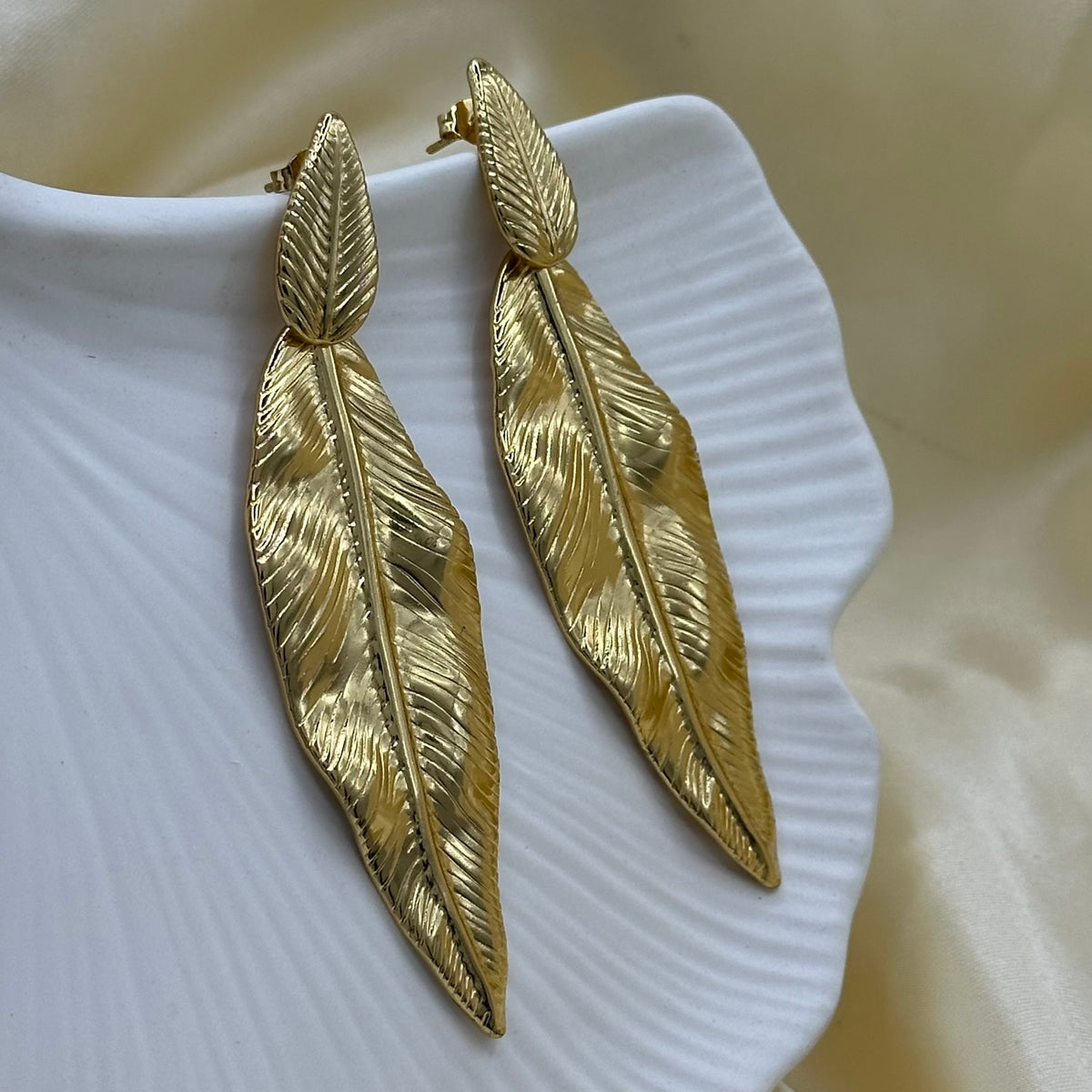 A pair of gold feather drop earrings with a textured, leaf-like design, displayed on a white scalloped dish against a soft beige satin background.
