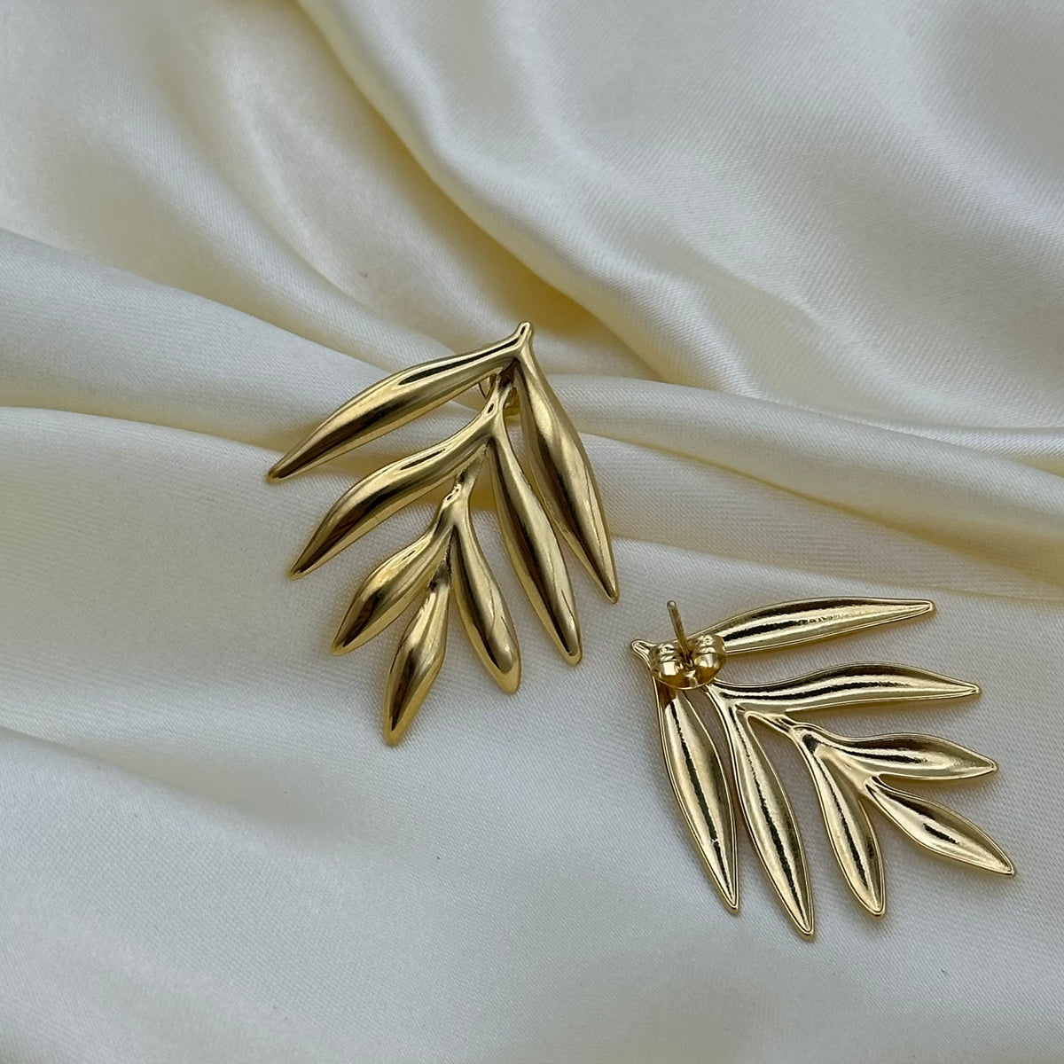 A pair of gold leaf-shaped stud earrings displayed on draped cream satin fabric.