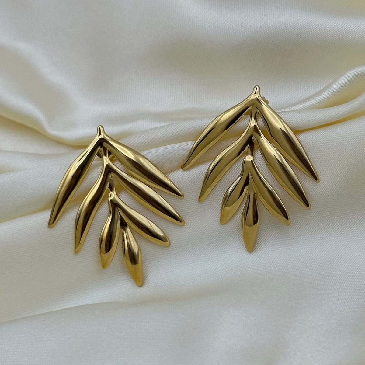 A pair of gold leaf-shaped stud earrings displayed on a draped cream satin fabric background.