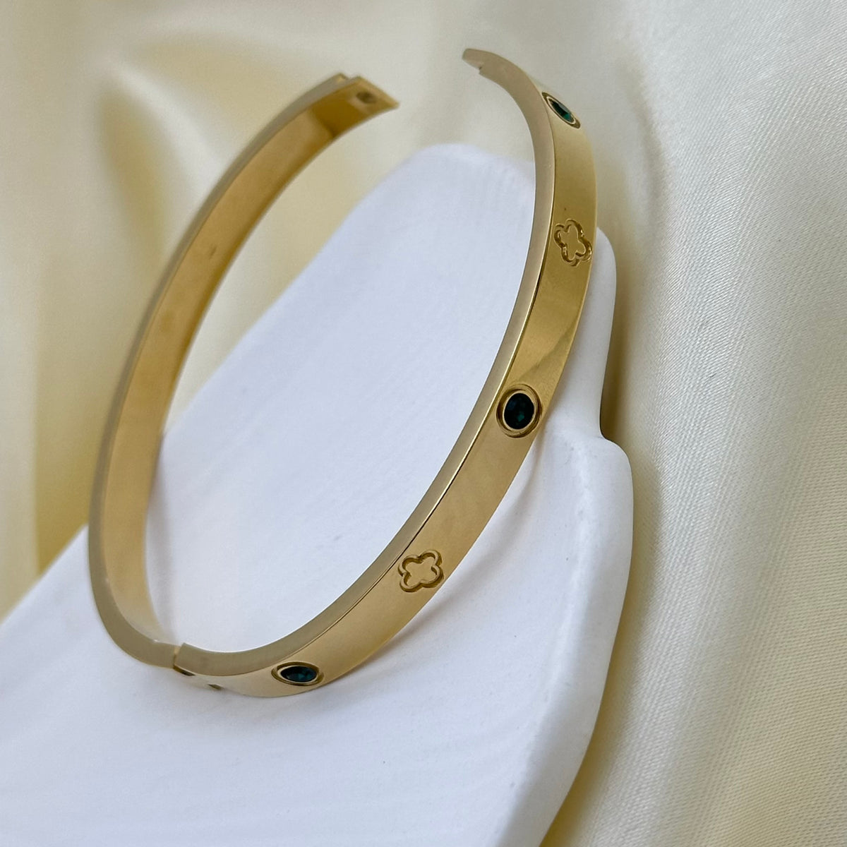 A gold-toned open cuff bangle bracelet with small green stone and clover-shaped inlays, displayed on a white stand against a soft cream fabric background.