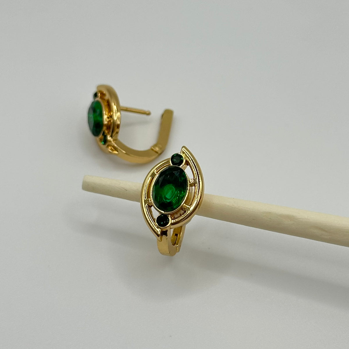A pair of gold hoop earrings with oval green gemstones and small dark accent stones, displayed on a white stand against a light gray background.