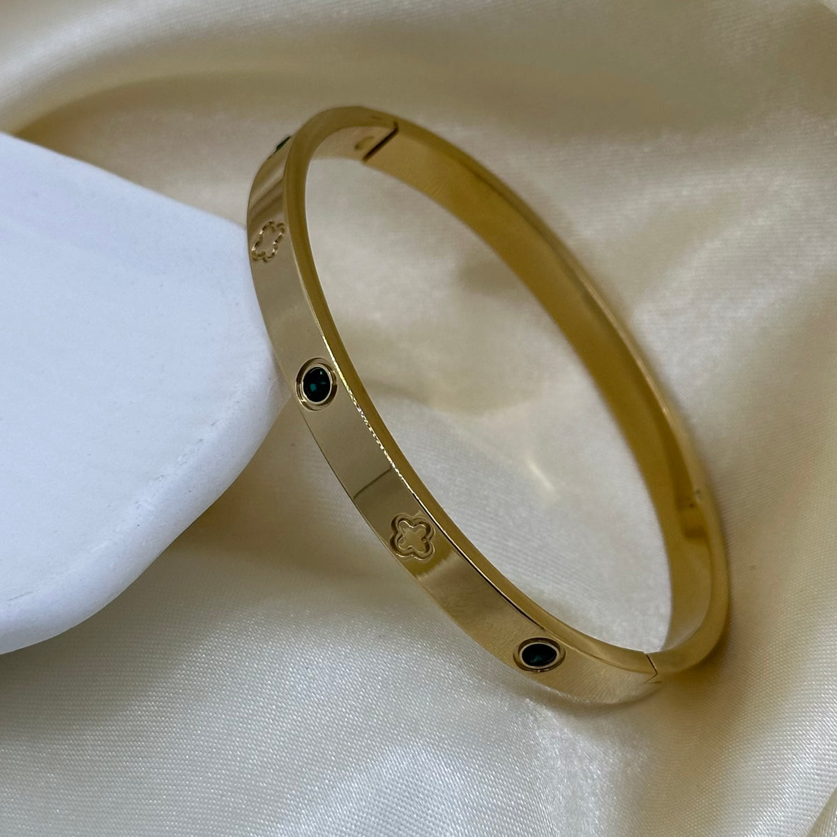 A gold bangle bracelet with small green circular accents and engraved clover motifs, resting on a cream satin fabric next to a white jewellery display block.