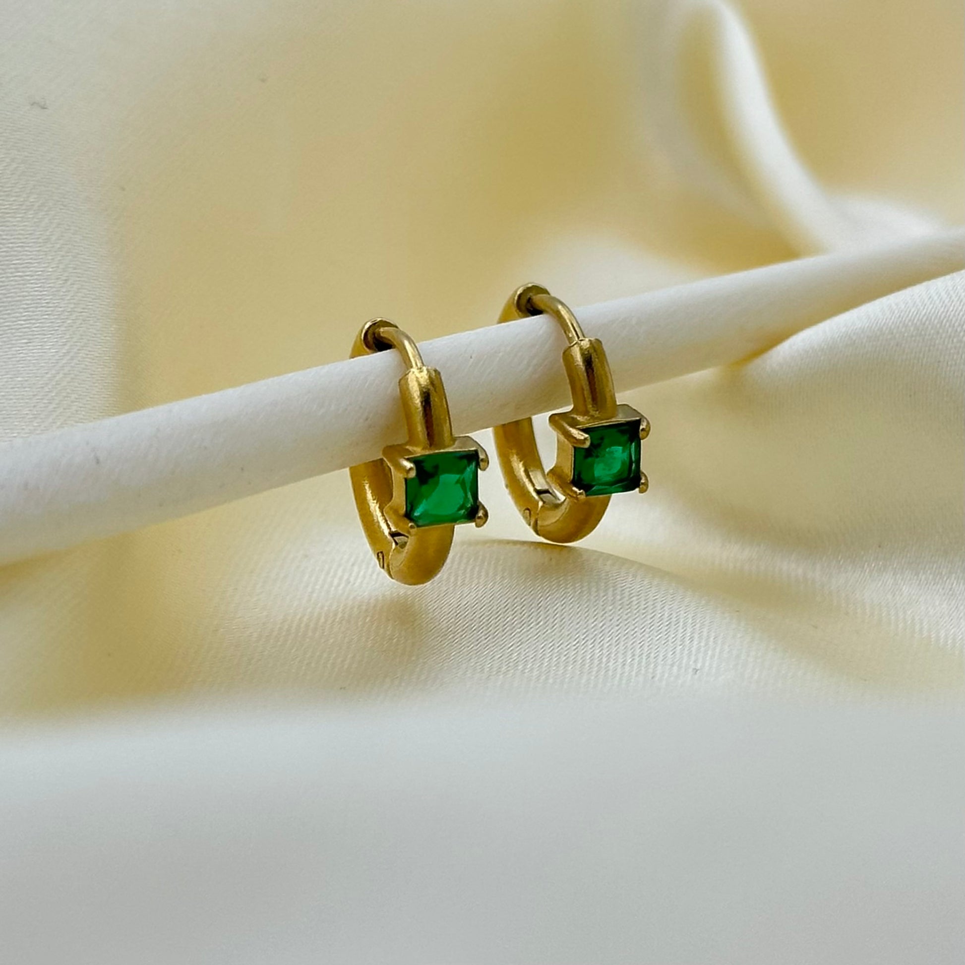 A pair of gold huggie hoop earrings with square green gemstones, displayed on a white stand against a soft cream-colored fabric background.