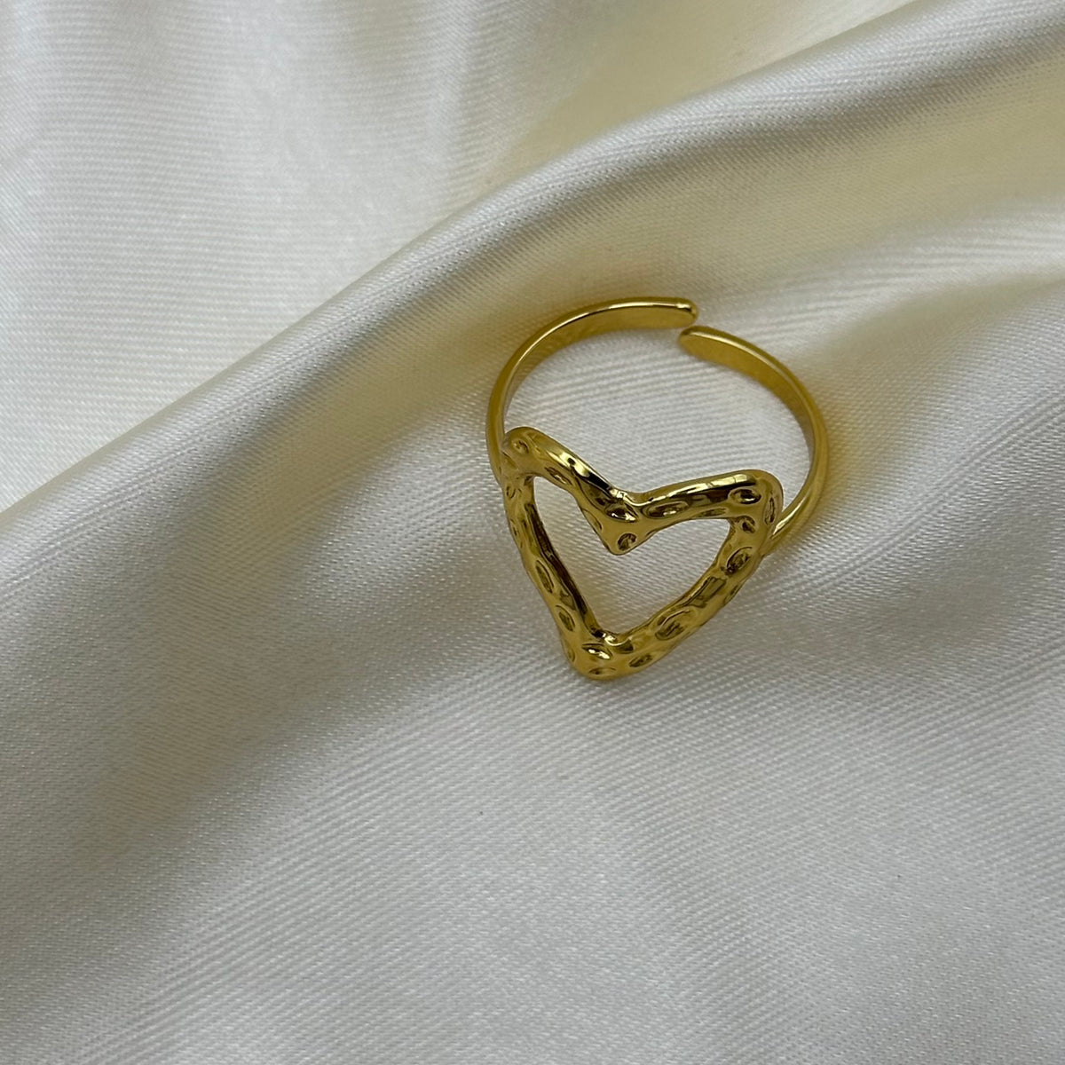 A gold heart-shaped open ring with a textured, dotted border design, resting on a draped cream-colored satin fabric.