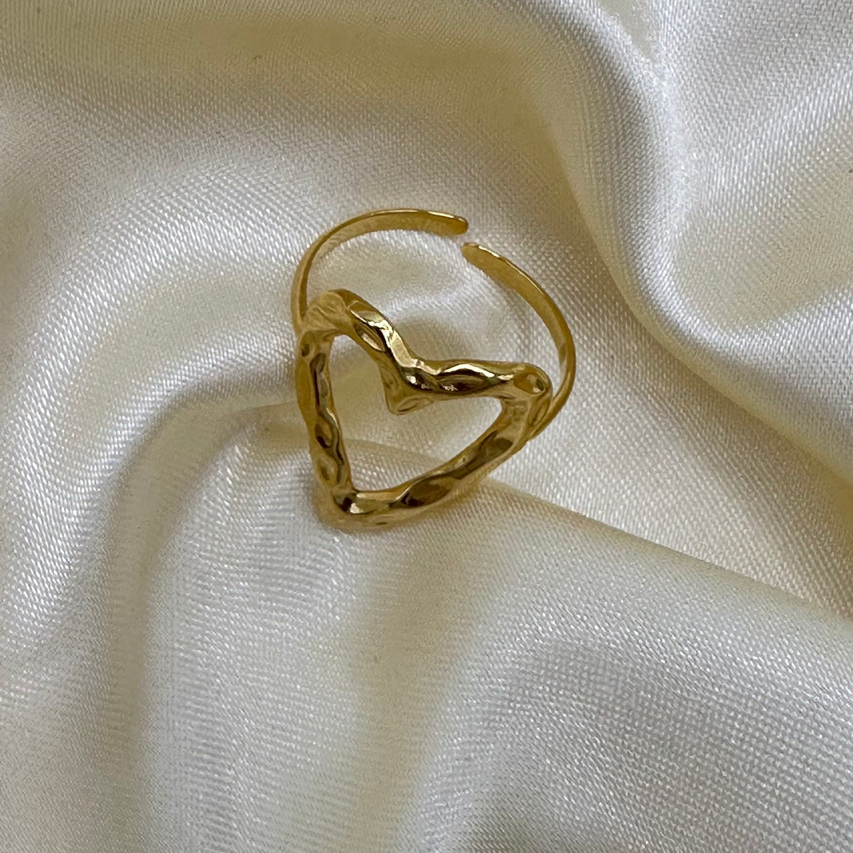 A hammered gold heart-shaped ring with an adjustable open band, displayed on white satin fabric.