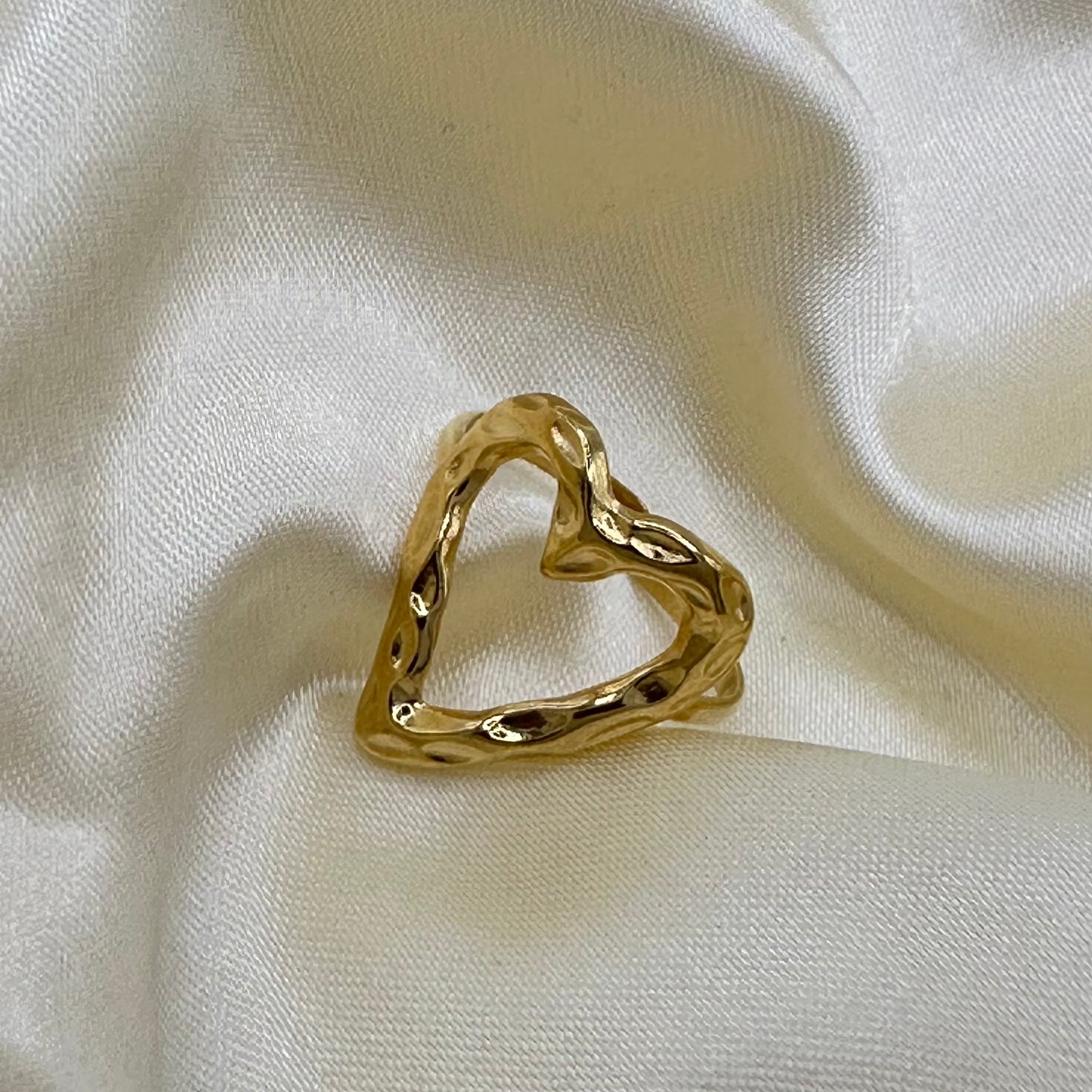 A hammered gold open-heart ring lying on draped white satin fabric.