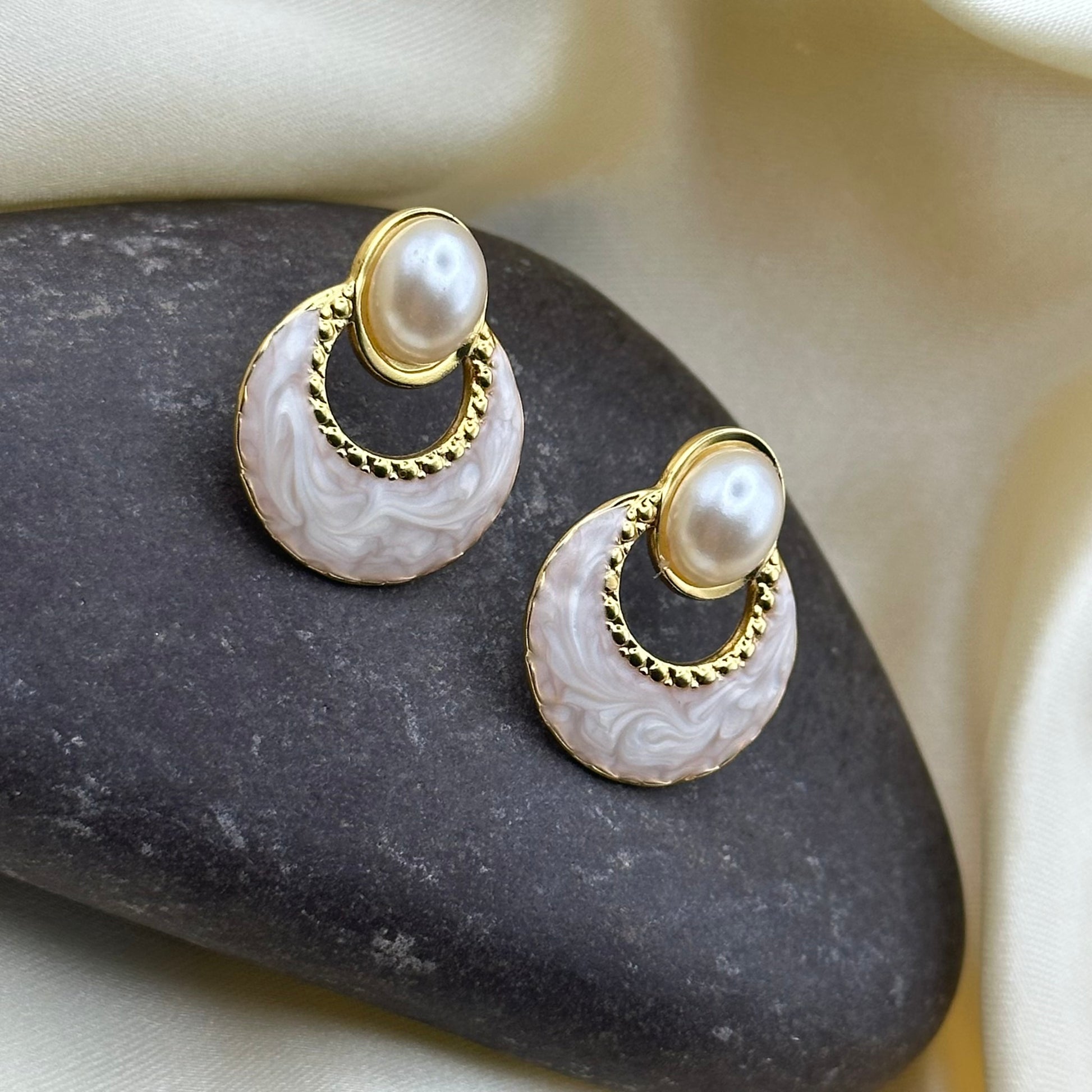 A pair of gold-tone earrings with oval faux pearls and pale grey marbled enamel crescents, displayed on a dark stone against cream satin fabric.