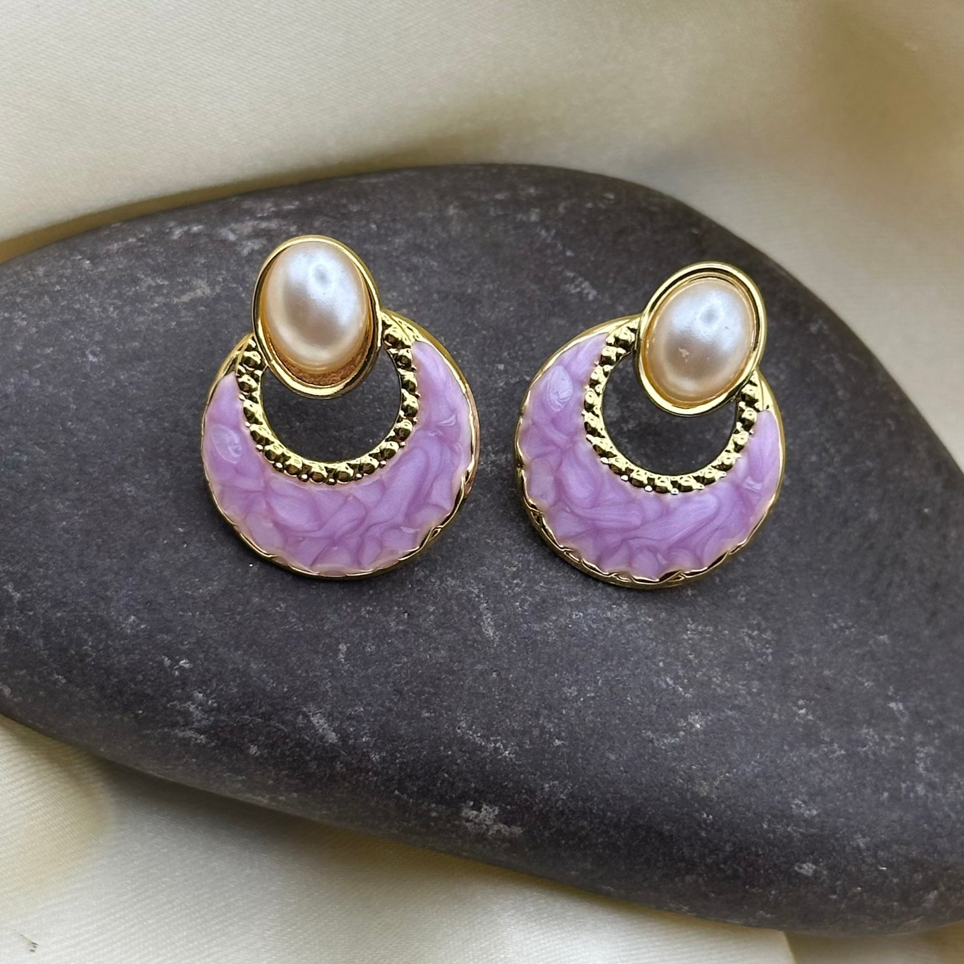 A pair of gold-tone earrings with oval faux pearls and lilac-pink enamel crescents, displayed on a dark stone against cream satin fabric.