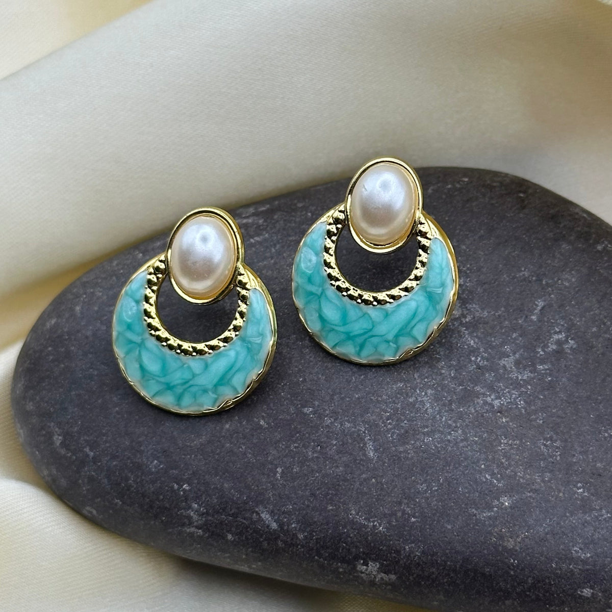 A pair of gold-tone earrings with oval faux pearls and turquoise marbled enamel crescents, displayed on a dark stone against cream satin fabric.