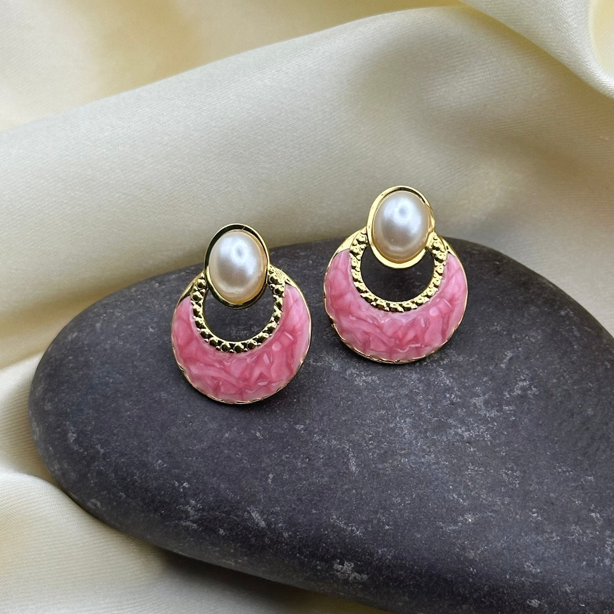 A pair of gold-tone earrings with oval faux pearls and rose-pink textured enamel crescents, displayed on a dark stone against cream satin fabric.