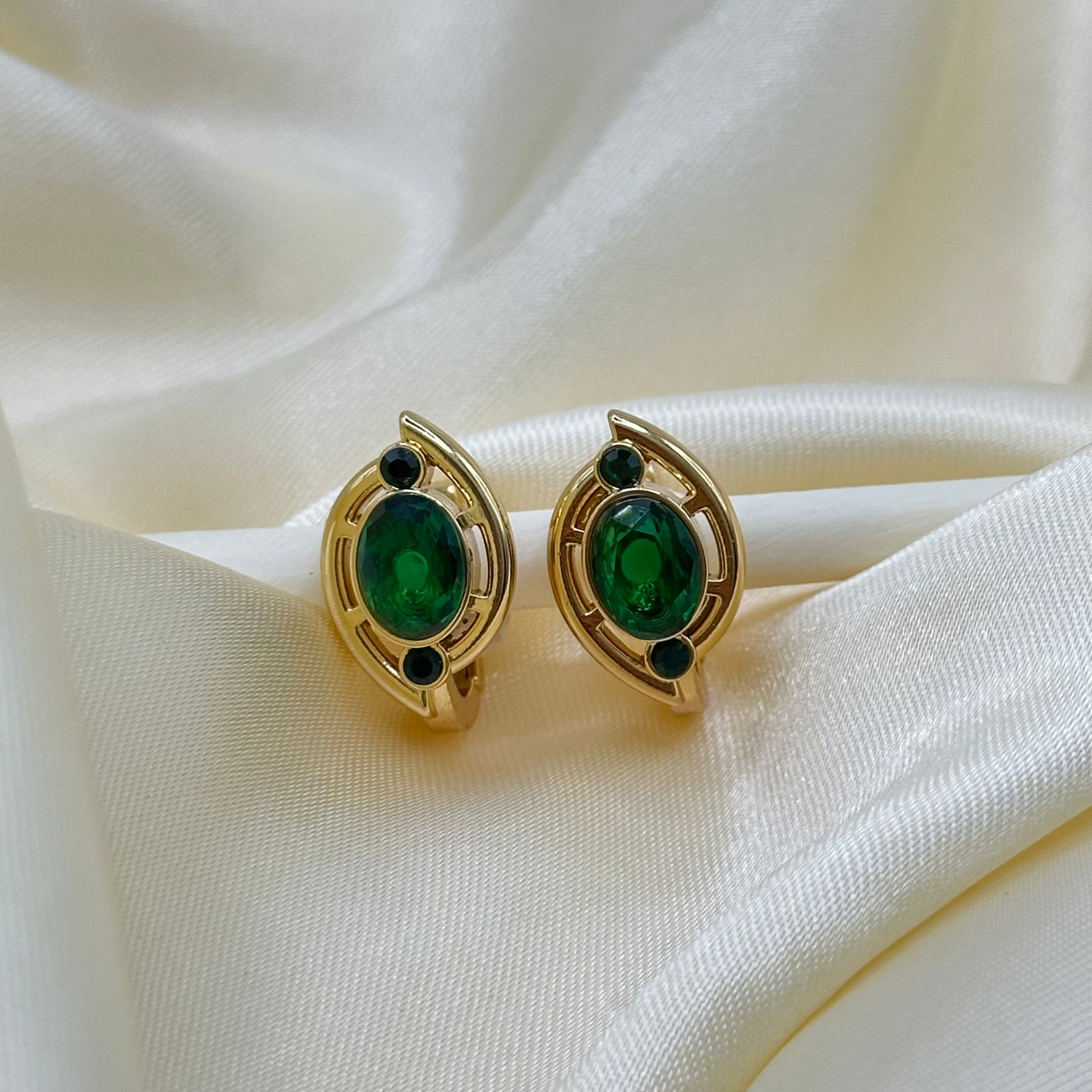 A pair of gold stud earrings with oval emerald-green gemstones and small dark accent stones, displayed on draped cream satin fabric.