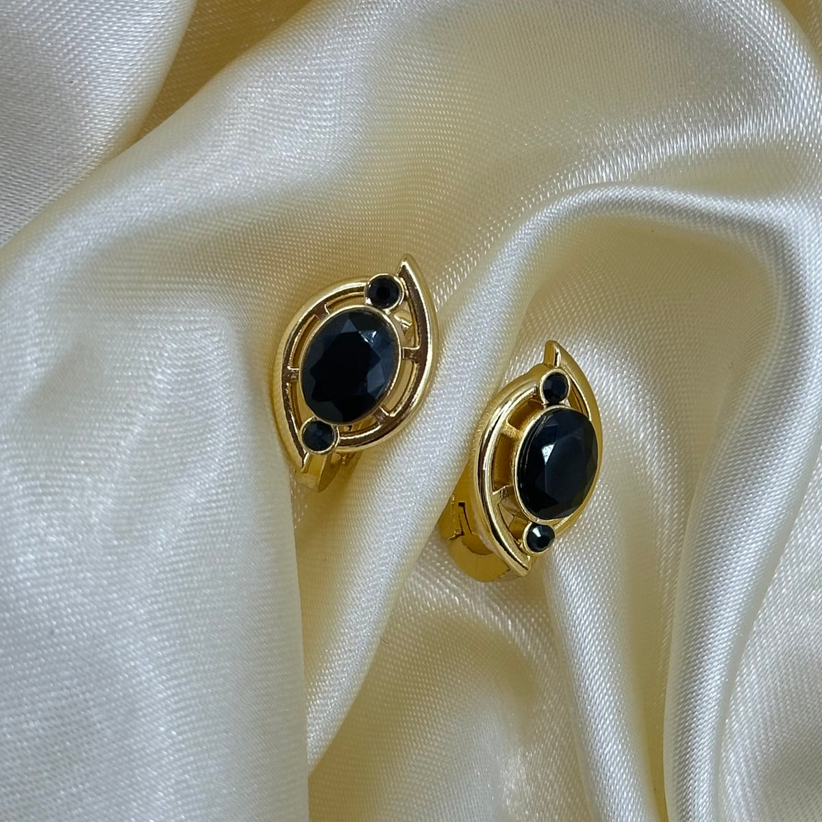 A pair of gold stud earrings with oval black gemstones and small black accent stones, displayed on draped cream satin fabric.