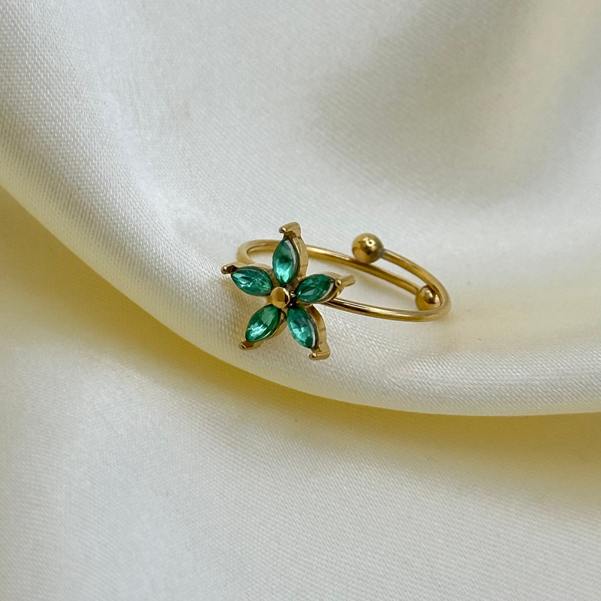 A gold adjustable ring with an emerald-green marquise-cut crystal flower, displayed on draped cream satin fabric.