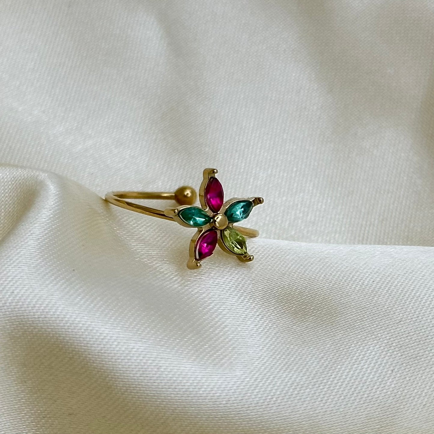A gold adjustable ring with a multicoloured crystal flower in pink, green and peridot tones, resting on cream satin fabric.