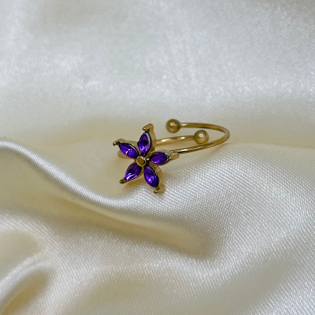 A gold adjustable ring with a purple marquise-cut crystal flower, displayed on draped cream satin fabric.