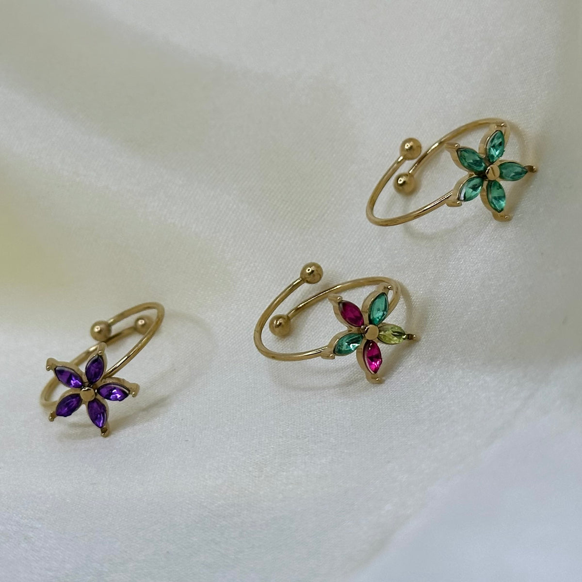 Three gold adjustable flower rings with purple, green and multicoloured crystal petals, arranged on soft cream fabric.