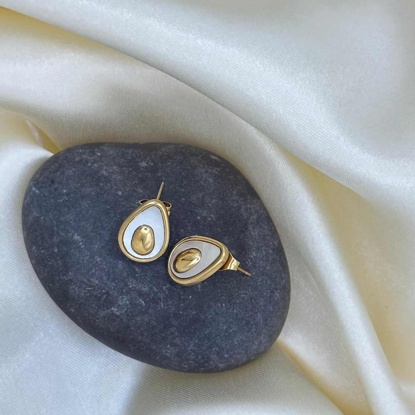 A pair of gold avocado-shaped stud earrings with white enamel and a polished gold pit centre, displayed on a dark stone over cream satin fabric.