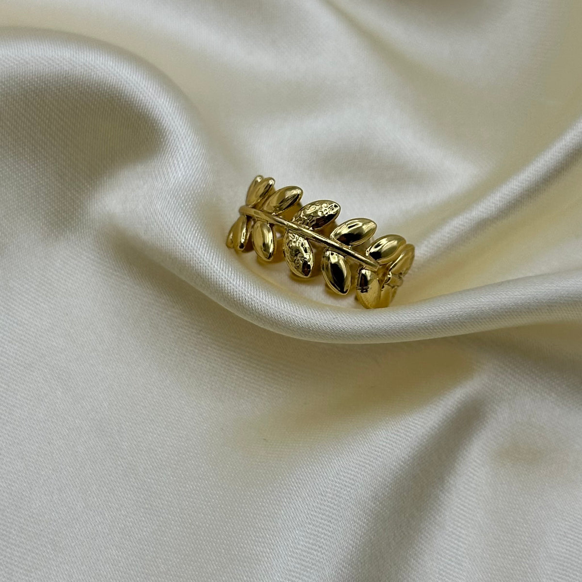 A gold laurel leaf band ring lying flat on cream satin, showing its detailed leaf pattern.