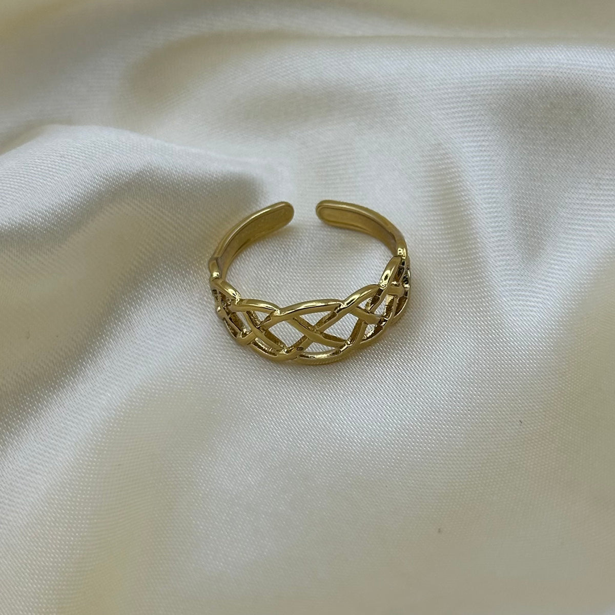 A gold-toned open cuff ring with an intricate woven or braided link pattern, resting on a draped cream-colored fabric background.