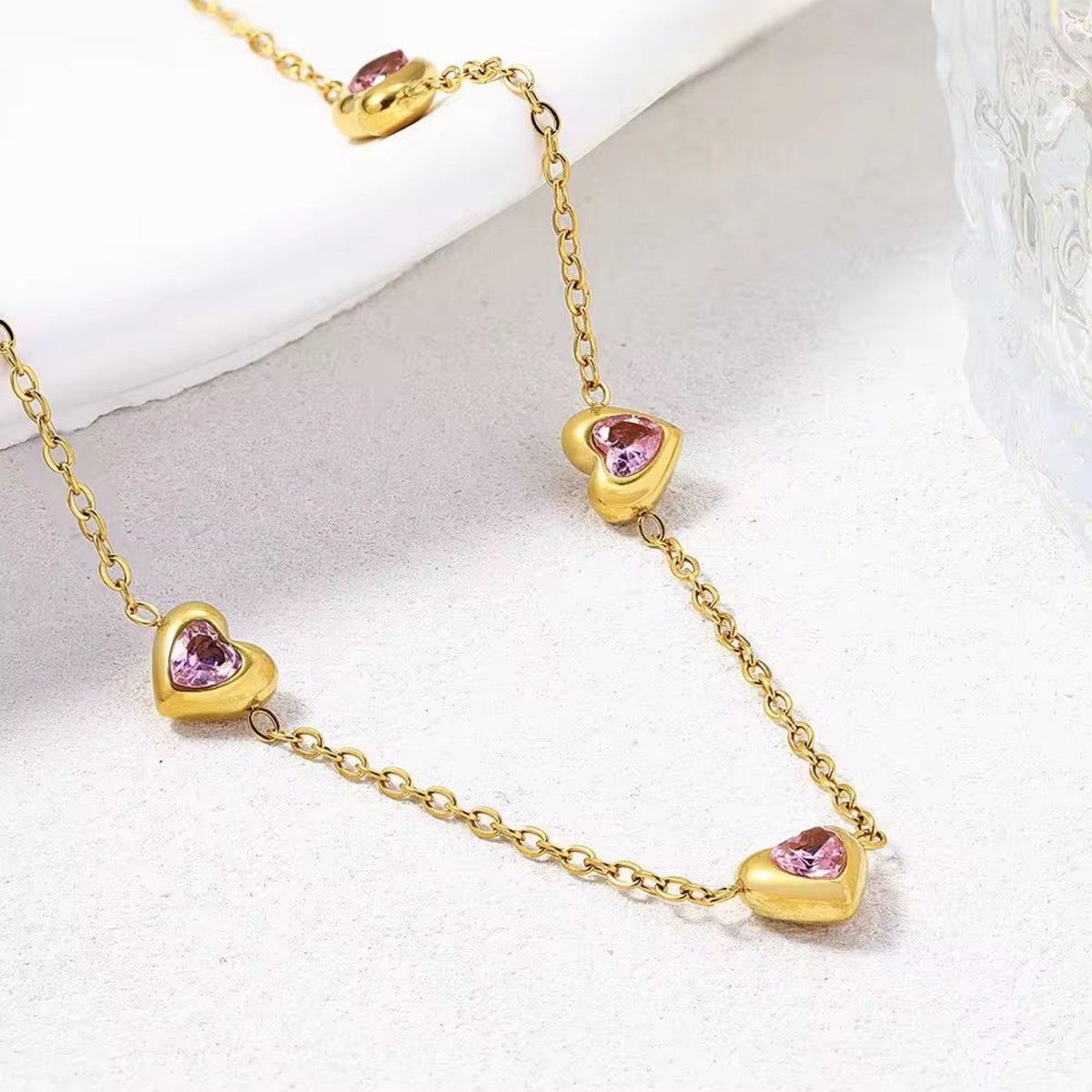 A gold chain necklace featuring heart-shaped charms, each inlaid with a small pink heart-shaped gemstone, displayed on a light textured surface.