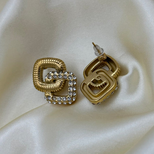 A pair of gold-tone geometric stud earrings with rhinestone accents, displayed on a draped cream satin fabric background.