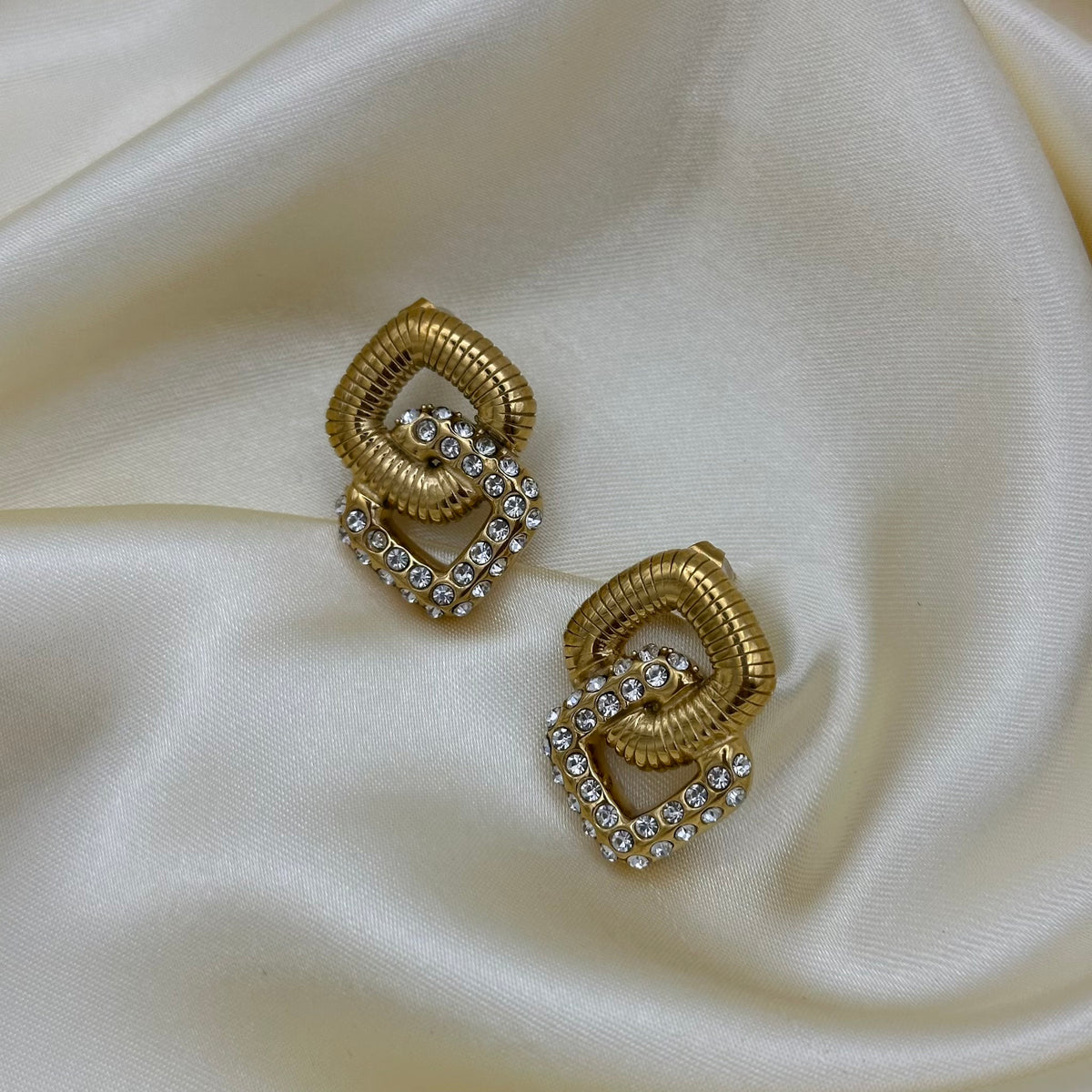 A pair of gold geometric stud earrings with clear rhinestones, displayed on a draped cream satin fabric background.