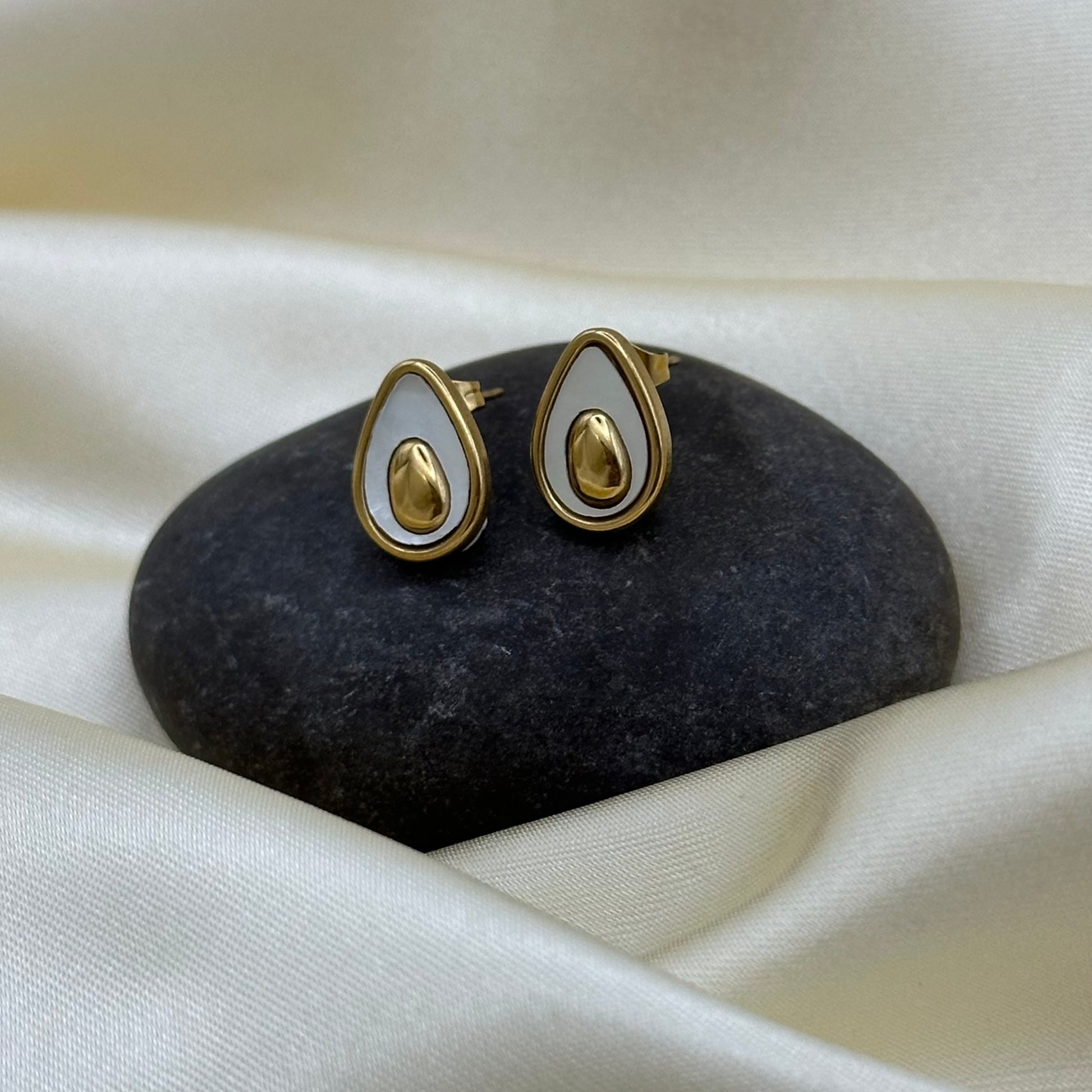 A pair of gold and white teardrop-shaped stud earrings displayed on a dark stone against a draped cream satin fabric background.