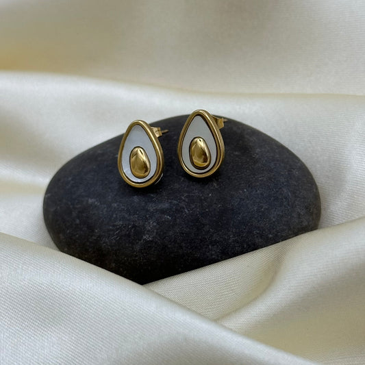 A pair of gold and white teardrop-shaped stud earrings displayed on a dark stone against a draped cream satin fabric background.