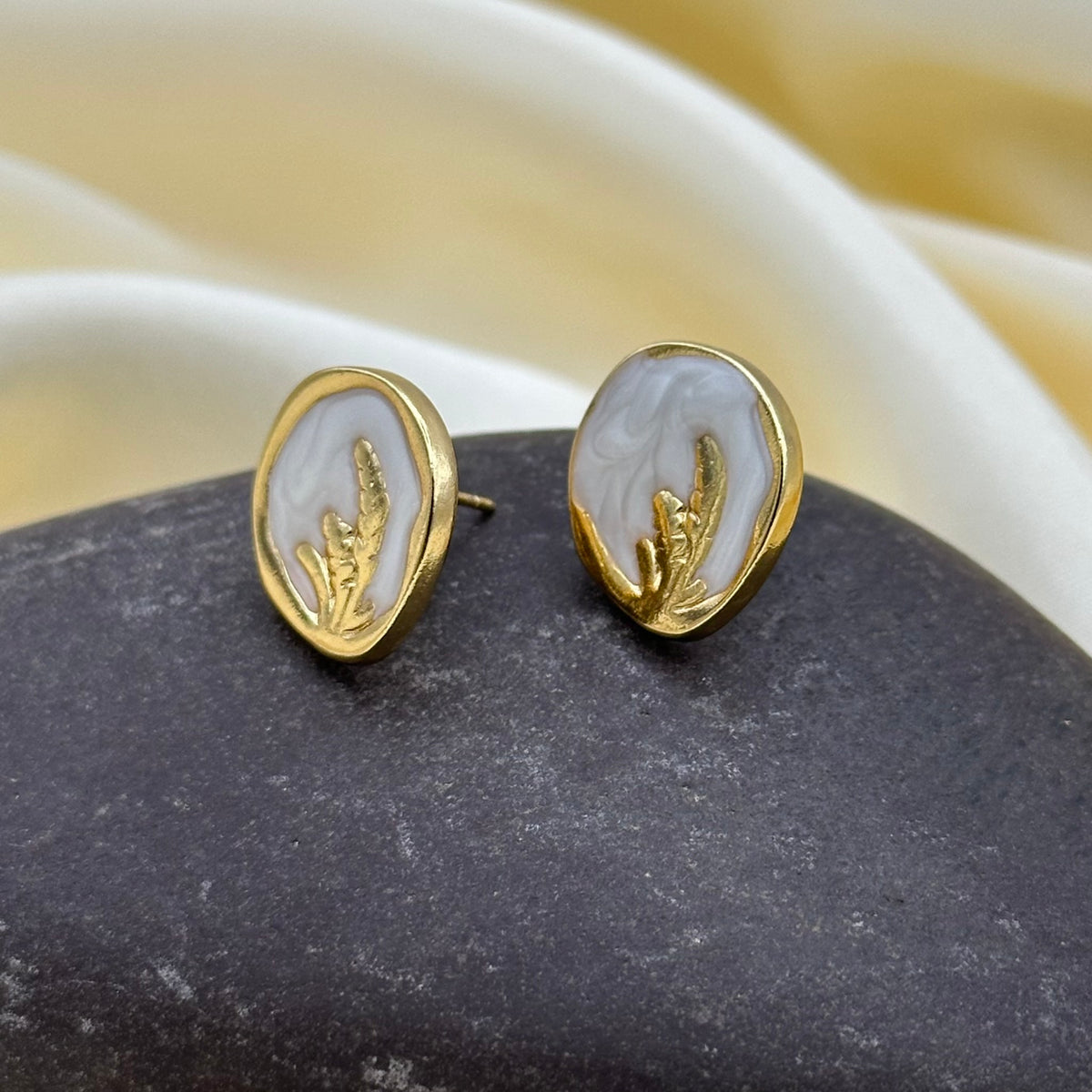 A pair of gold oval stud earrings with white inlays featuring a gold leaf design, displayed on a dark stone with a cream satin background.